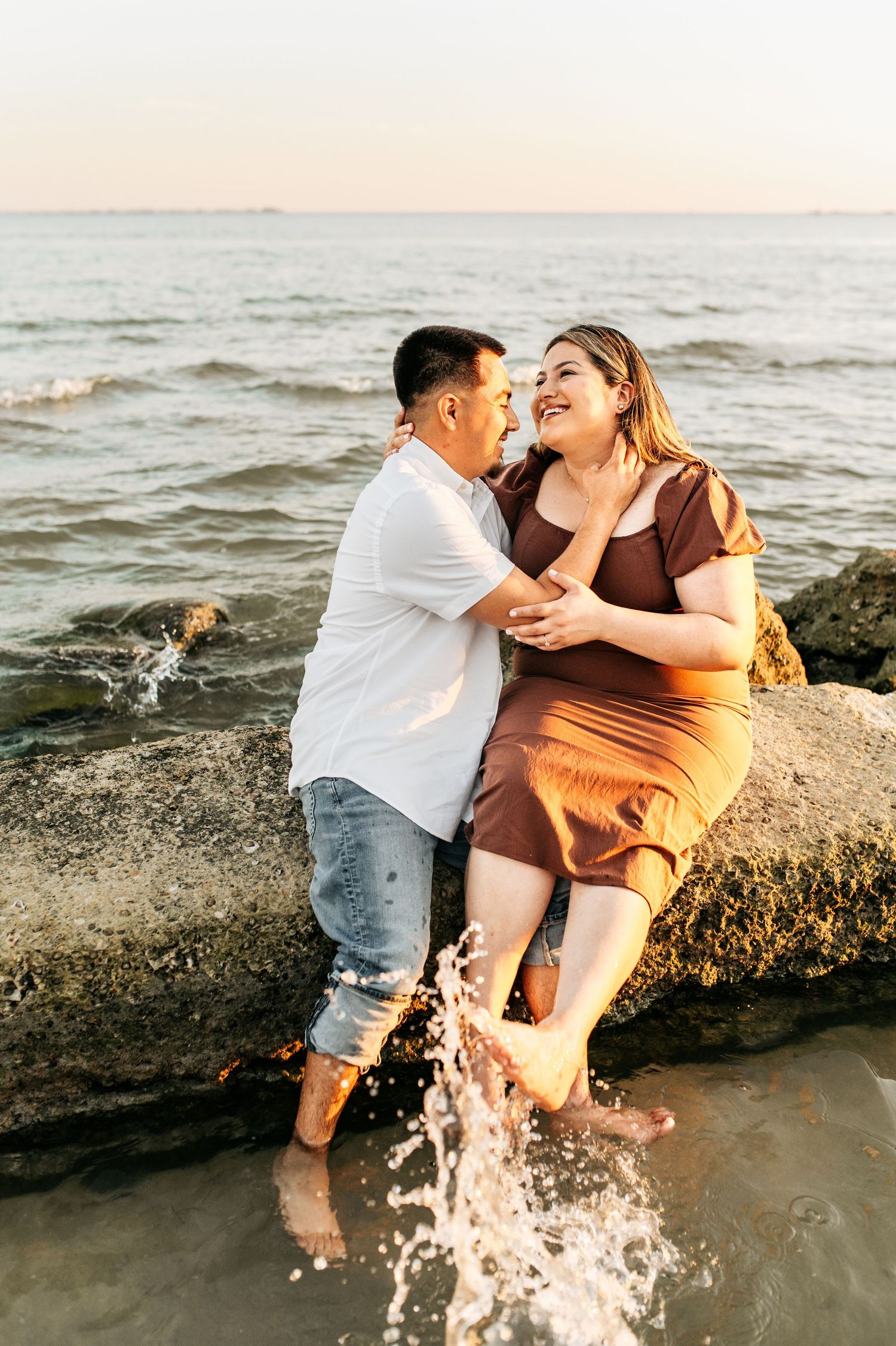 Playful couple splashing water and laughing together on rocky beach at sunset.