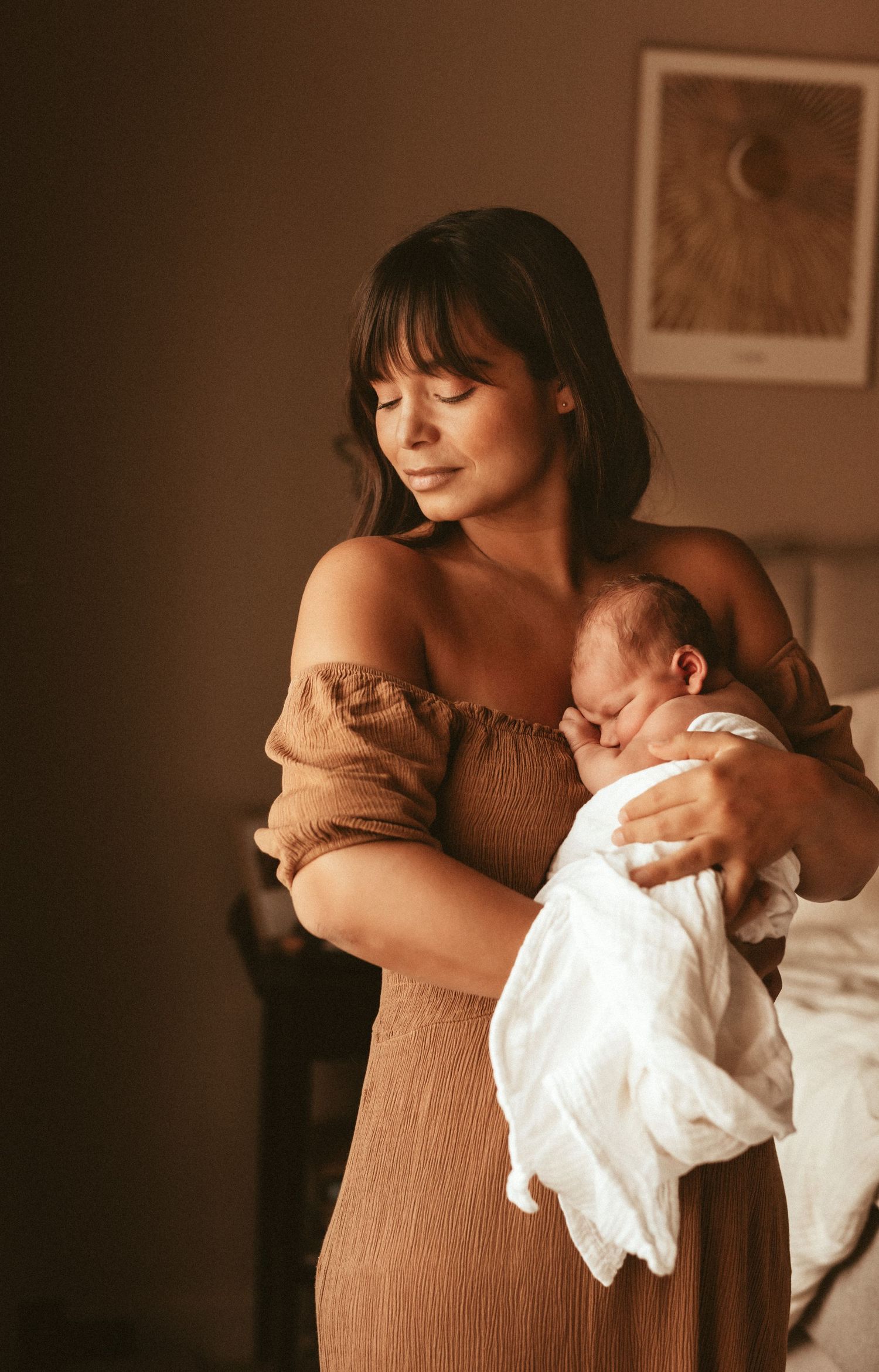 A mother in a brown dress tenderly holds her sleeping newborn baby wrapped in white in a dimly lit room. Newborn photographer Javea