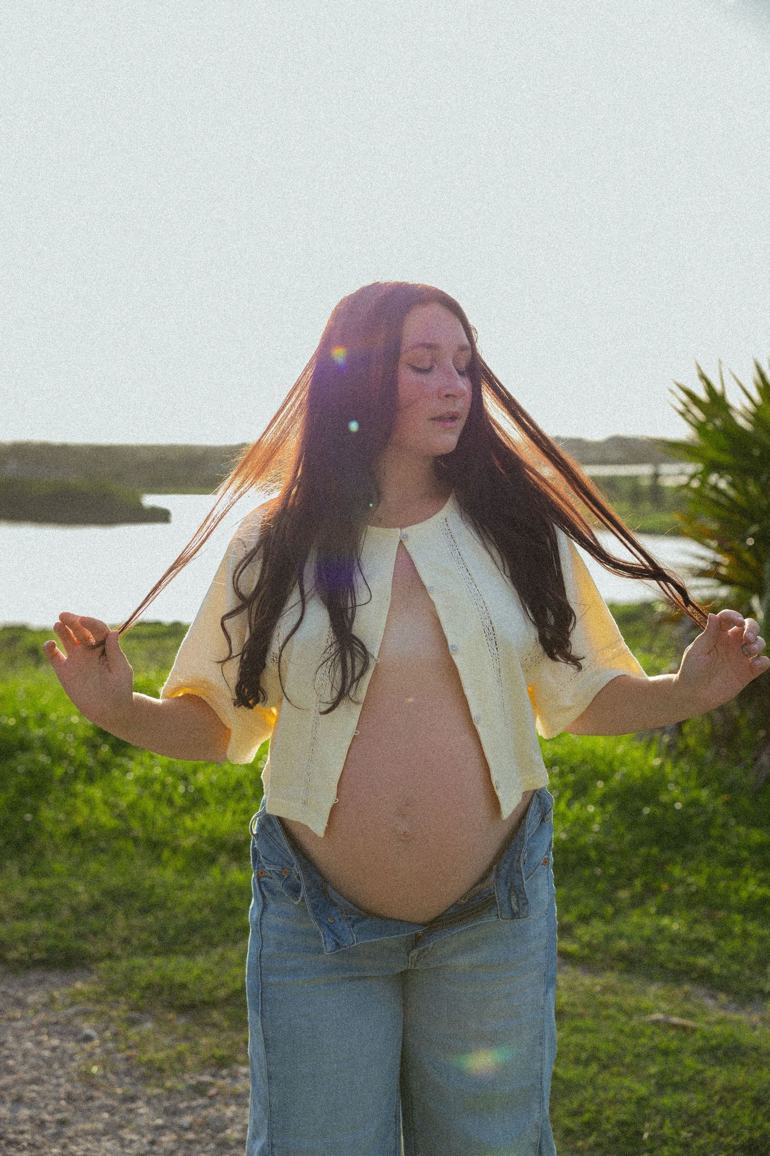 Person with long red hair poses expressively outdoors in jeans and white top at sunset by the water.