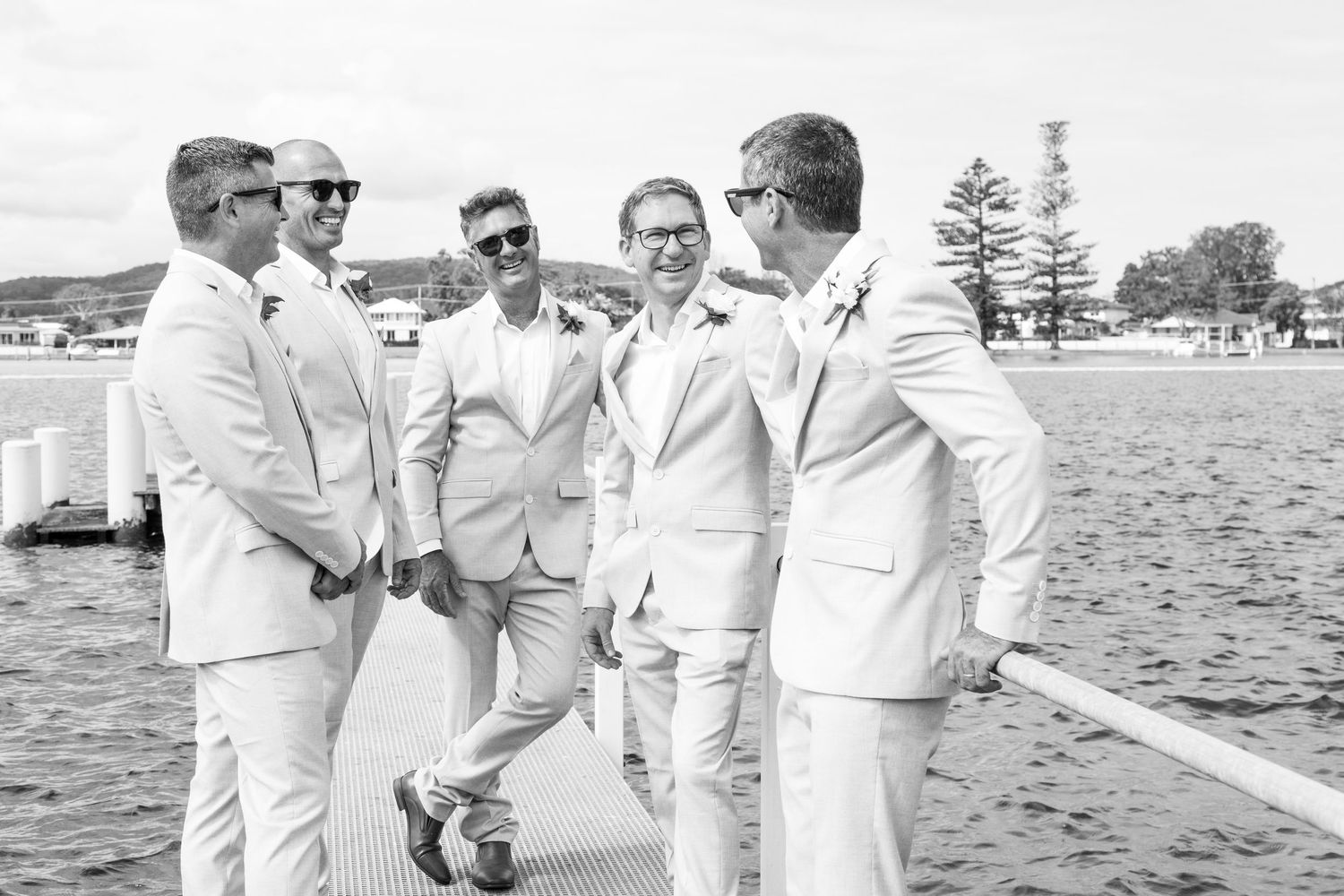 Groomsmen in matching white suits and sunglasses stand together chatting on a waterfront dock.