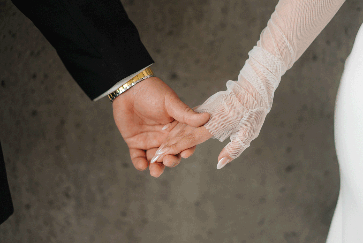 Two hands gently clasped together, one in a black sleeve and one in a white lace sleeve, symbolizing a wedding union.