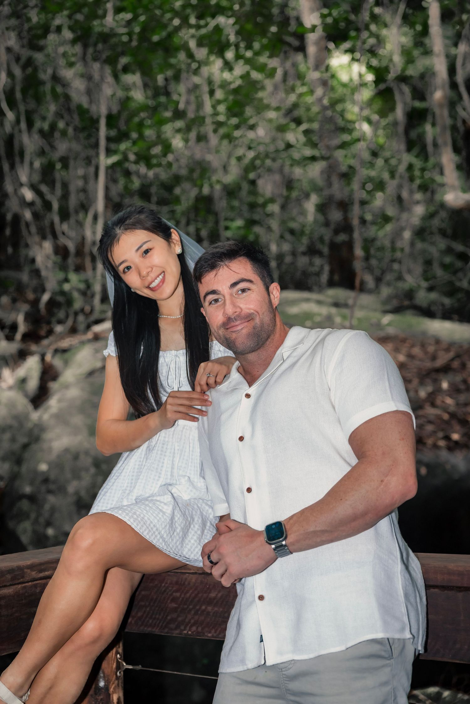 A couple dressed in white clothing sitting together on a rustic wooden bench in a wooded outdoor setting.