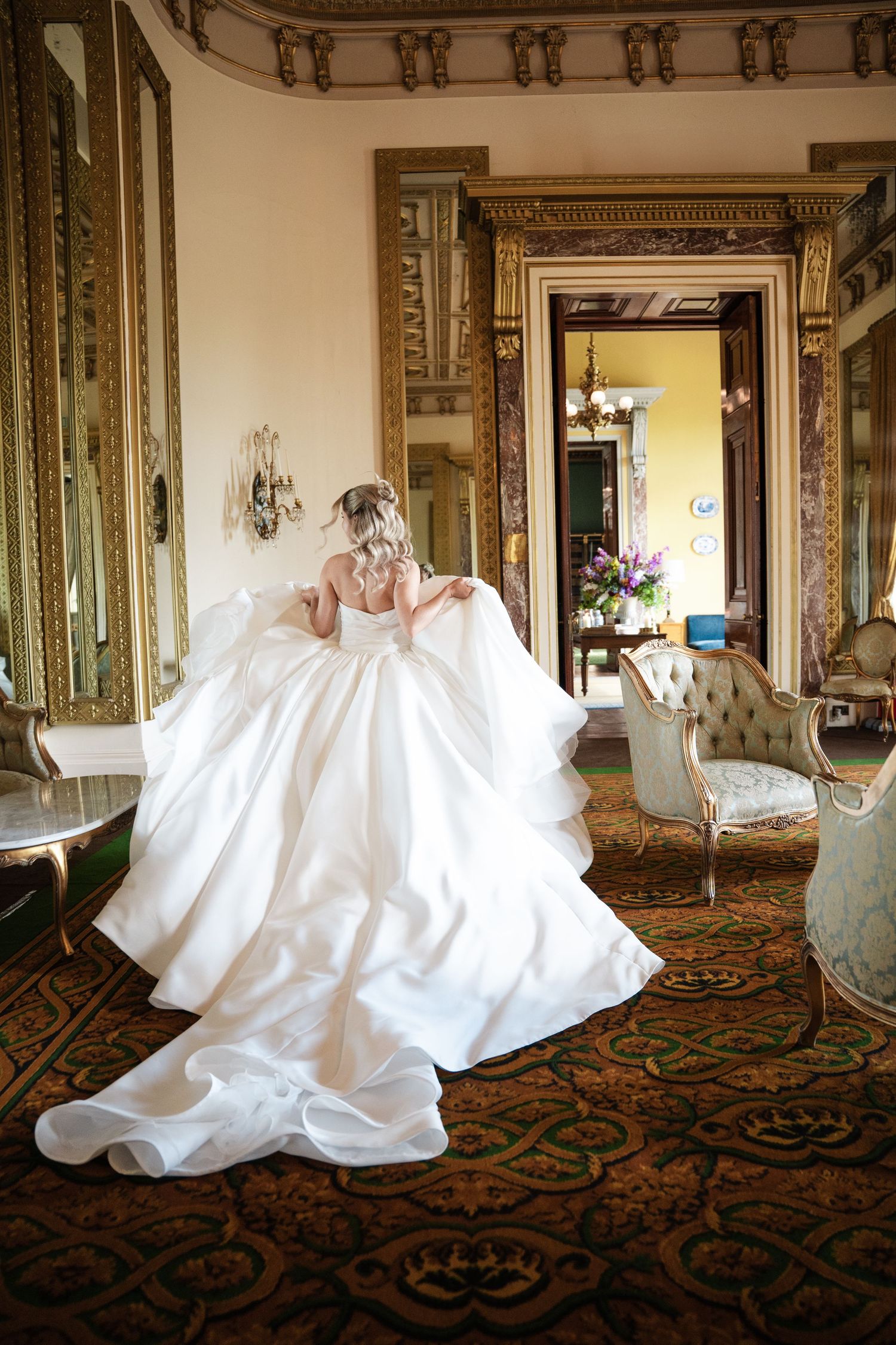 Elegant wedding dress with flowing train sweeps across ornate carpet in luxurious historic room with gilded mirrors and doorways.