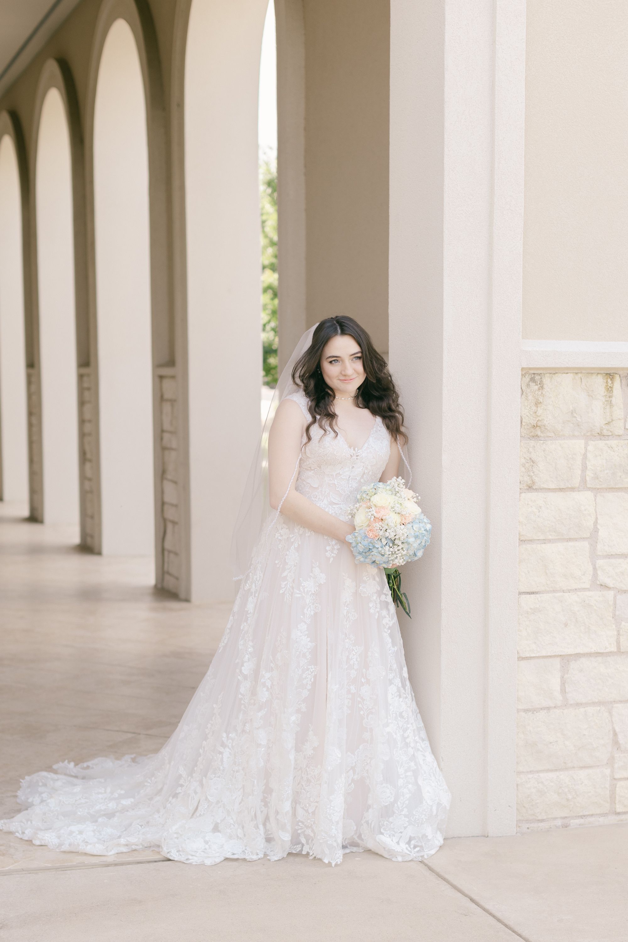Bride in lace wedding gown poses in elegant stone colonnade.