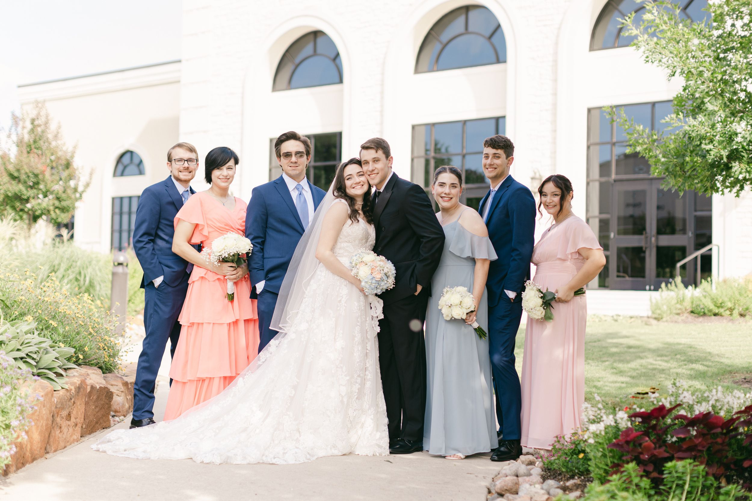 Wedding party in navy and coral attire poses outside elegant stone building.