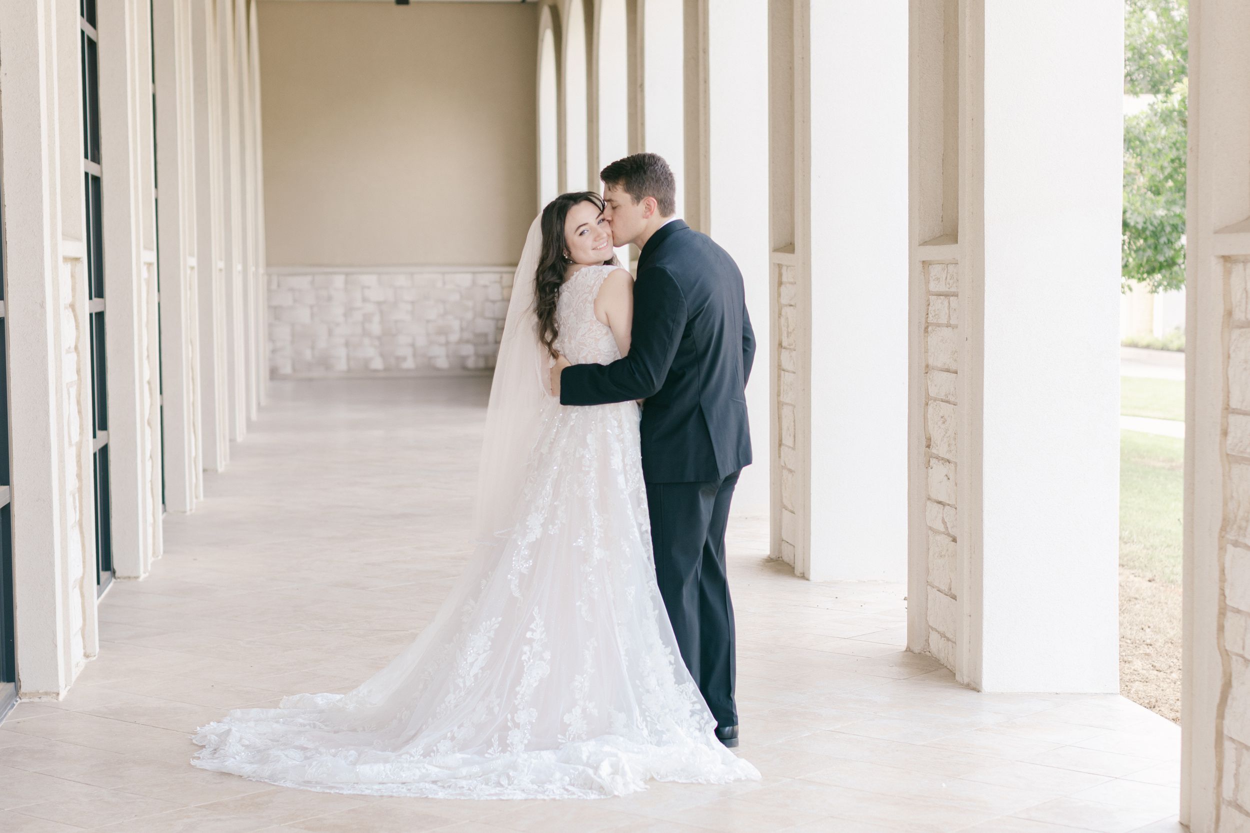 Elegant wedding couple poses in a bright white columned walkway with natural light streaming in.