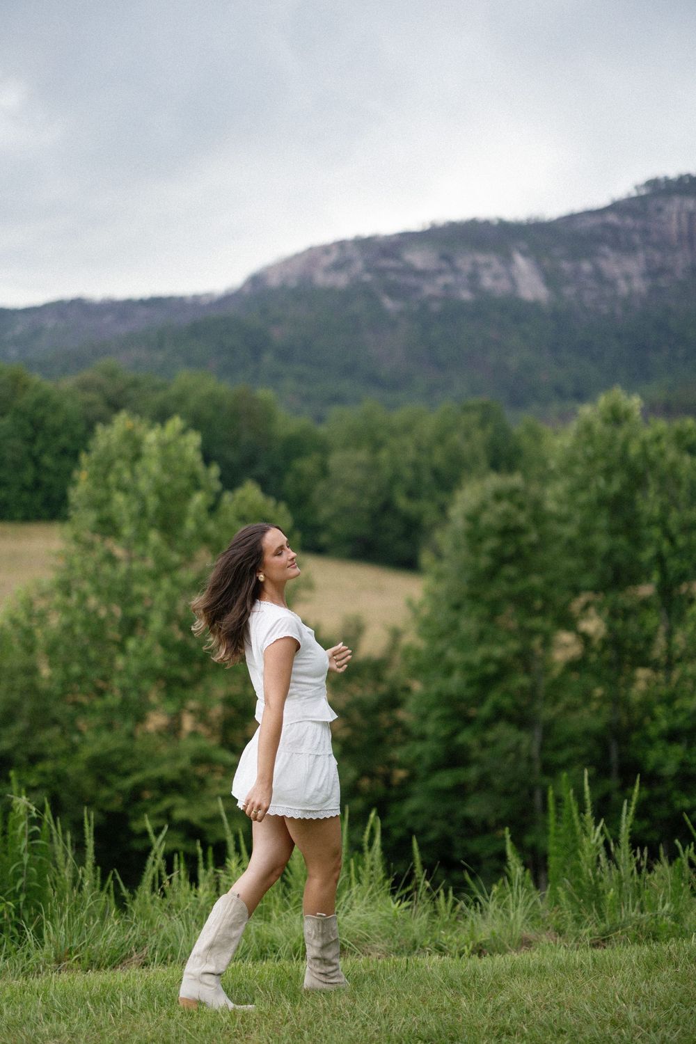 A person in a white dress and tall boots stands in a grassy field with mountains and trees in the background.