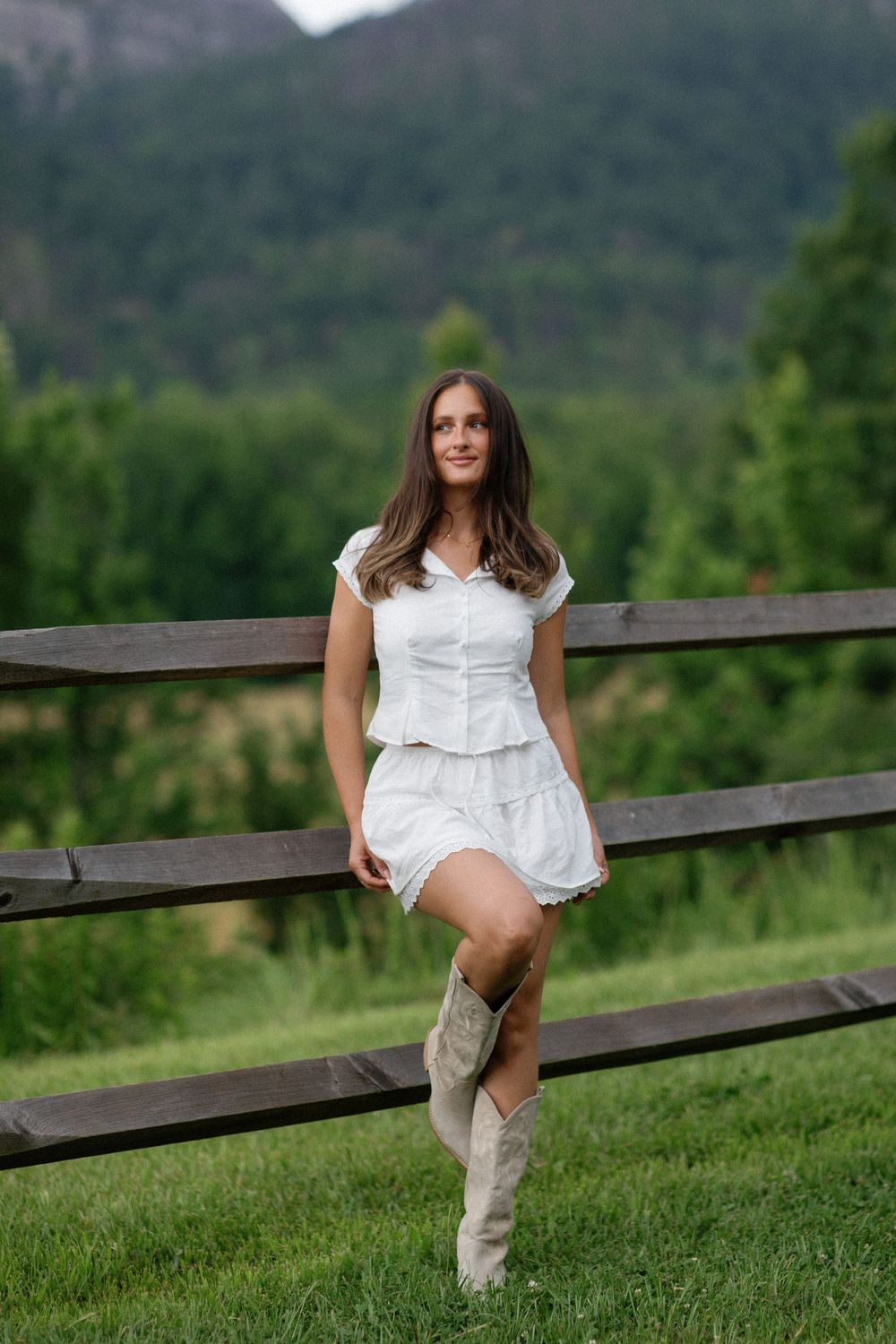 A woman in a white summer dress and tall boots sits on a wooden fence with mountains and green forest in the background.