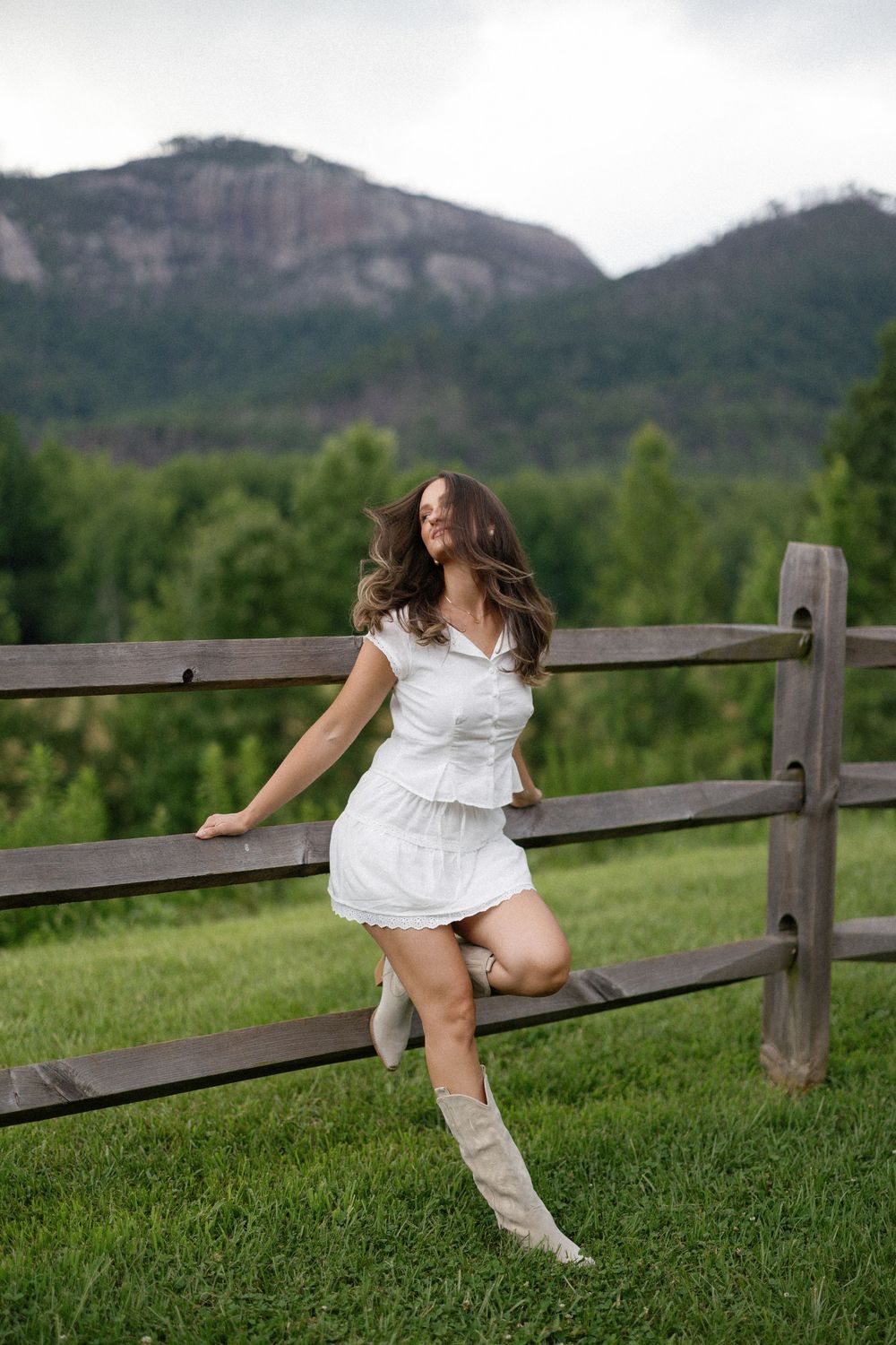 Person in a white dress and tall boots poses on a wooden fence with mountain scenery in the background.