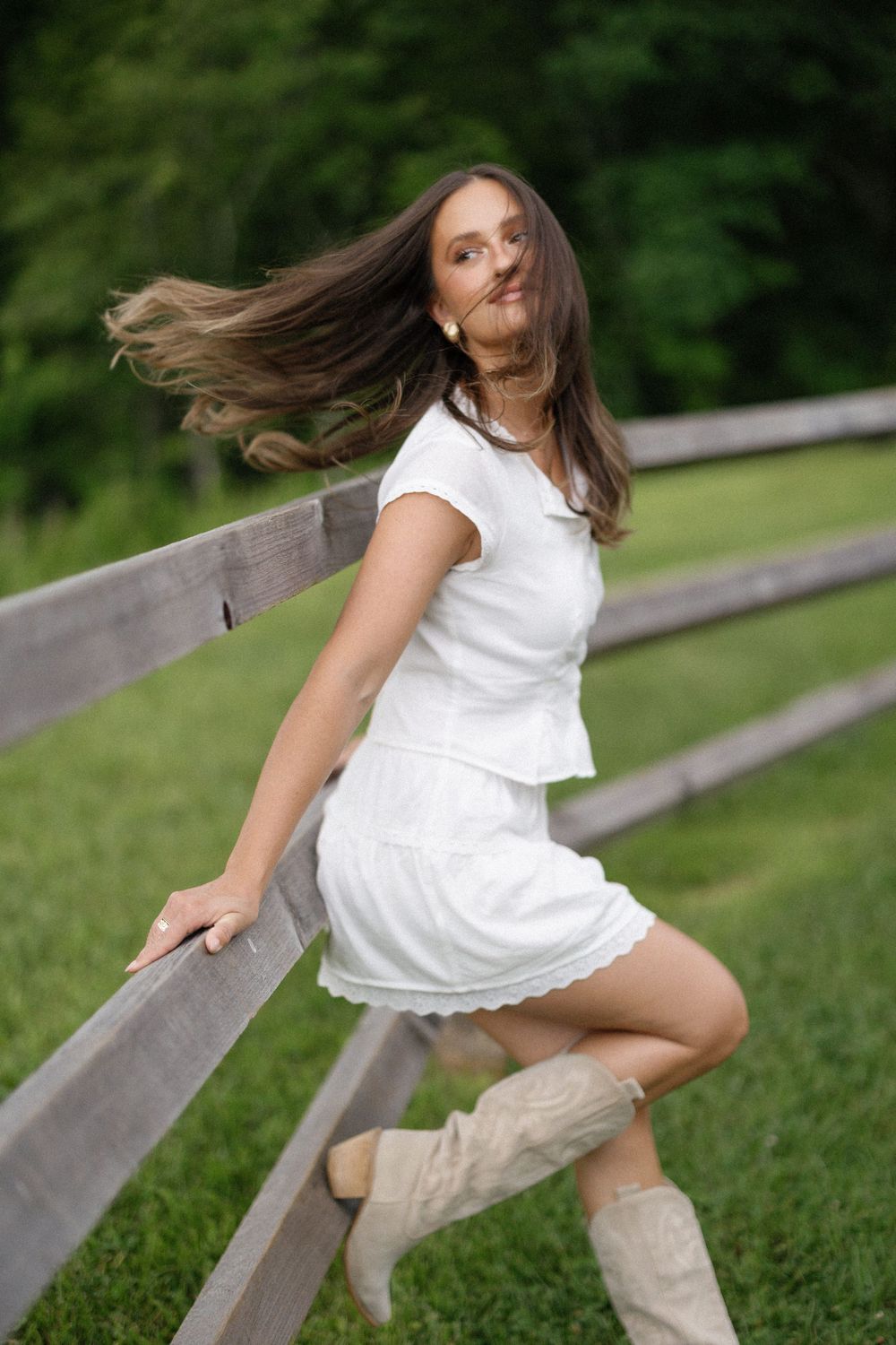 A figure in a white dress performs expressive dance movements while leaning on a wooden bench in a lush park setting.