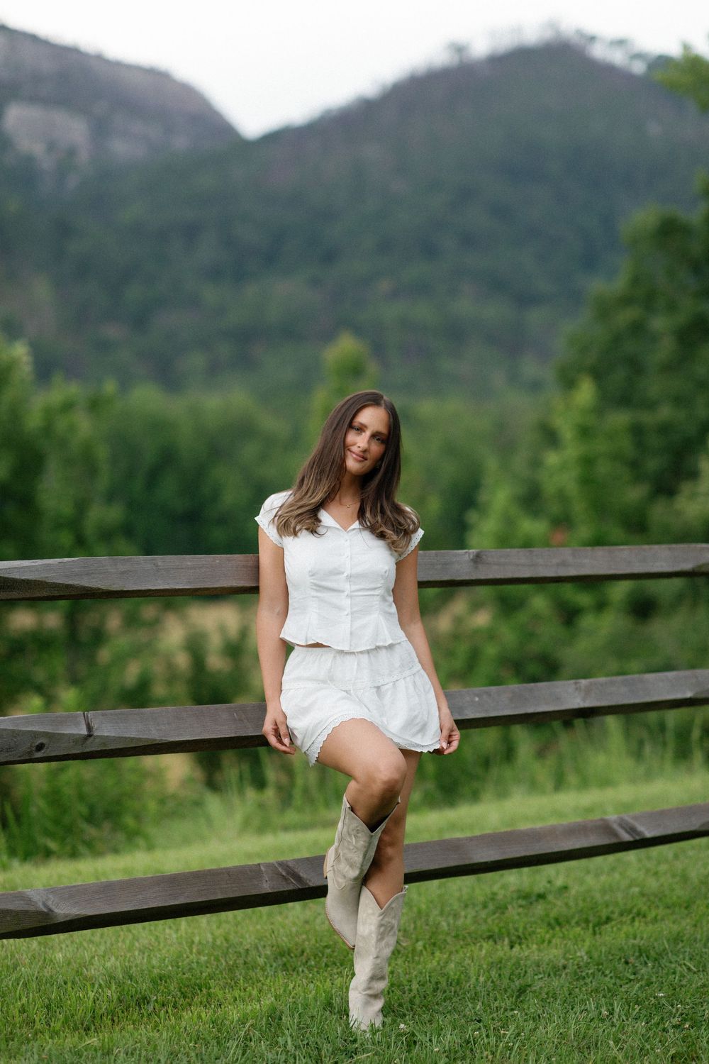 Country western style portrait with someone in a white dress and cowboy boots leaning on a wooden fence with mountains behind.