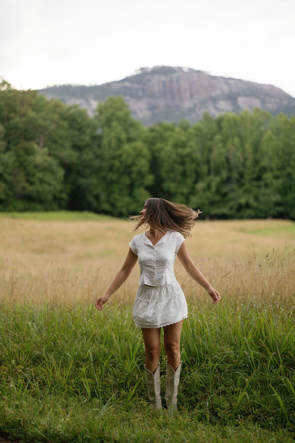 A person in a white dress spins in a golden field with tall boots, mountains and forest visible in the background.