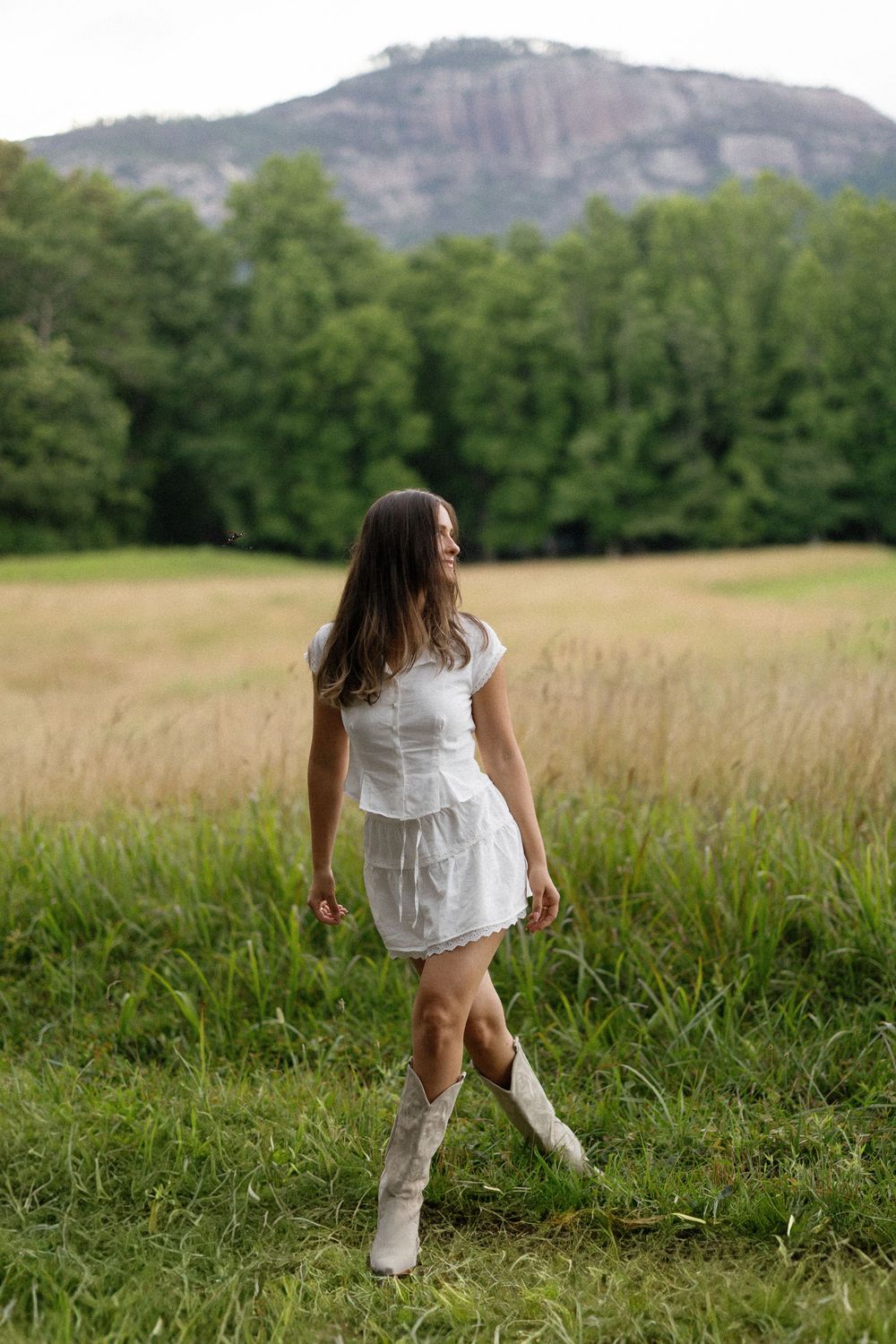 A series of five photos showing someone wearing a white dress playing in a lush green field with mountains in the background.