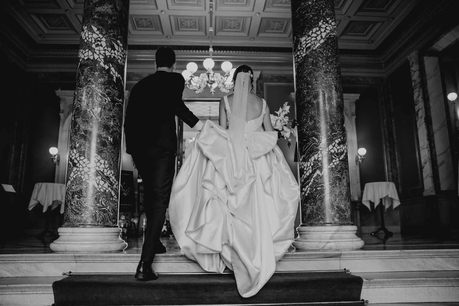 Two people in formal wedding attire walk through an ornate church interior with a grand coffered ceiling and columns.