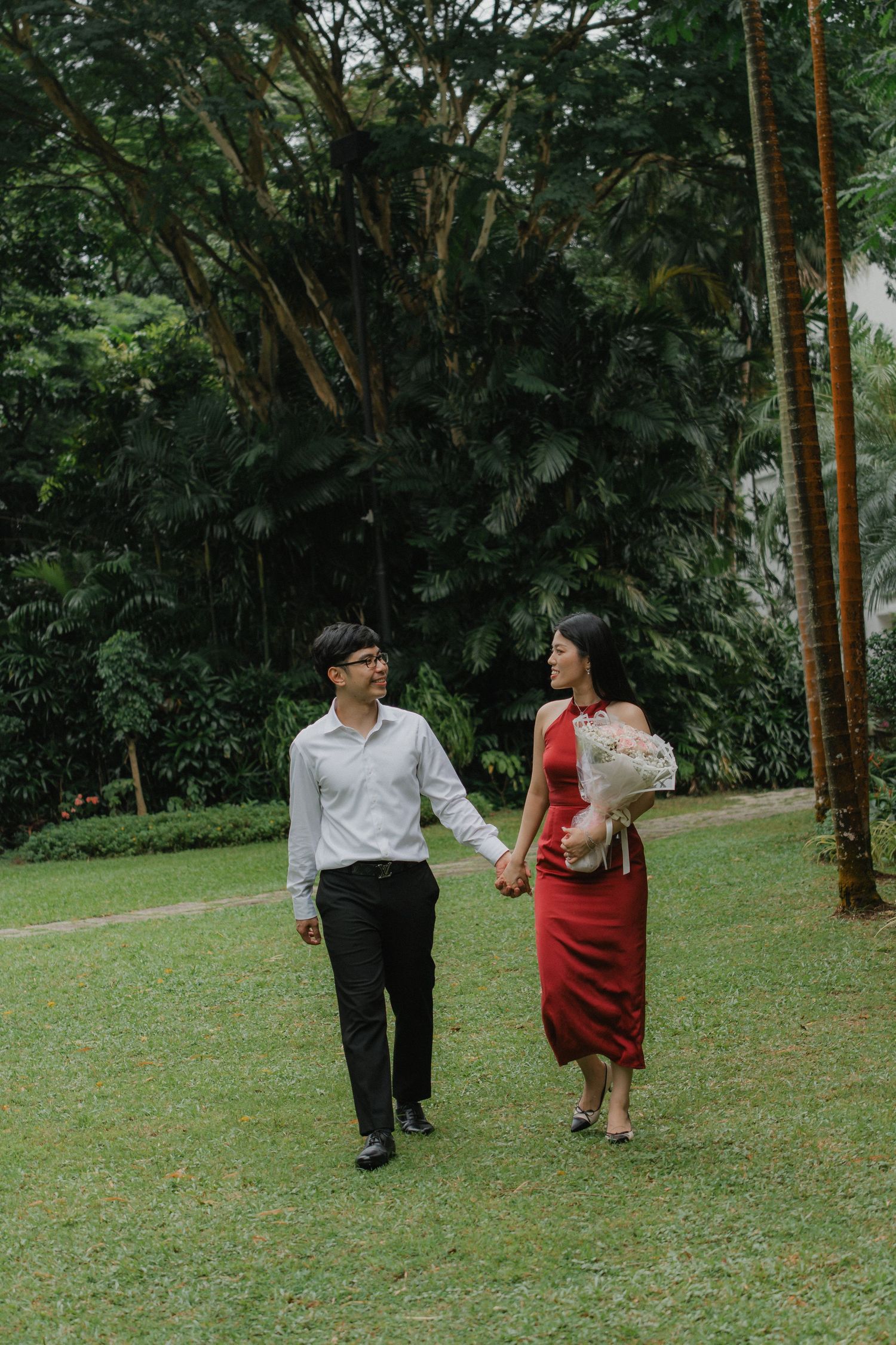 A couple strolls through a lush garden setting with tall trees and green grass.
