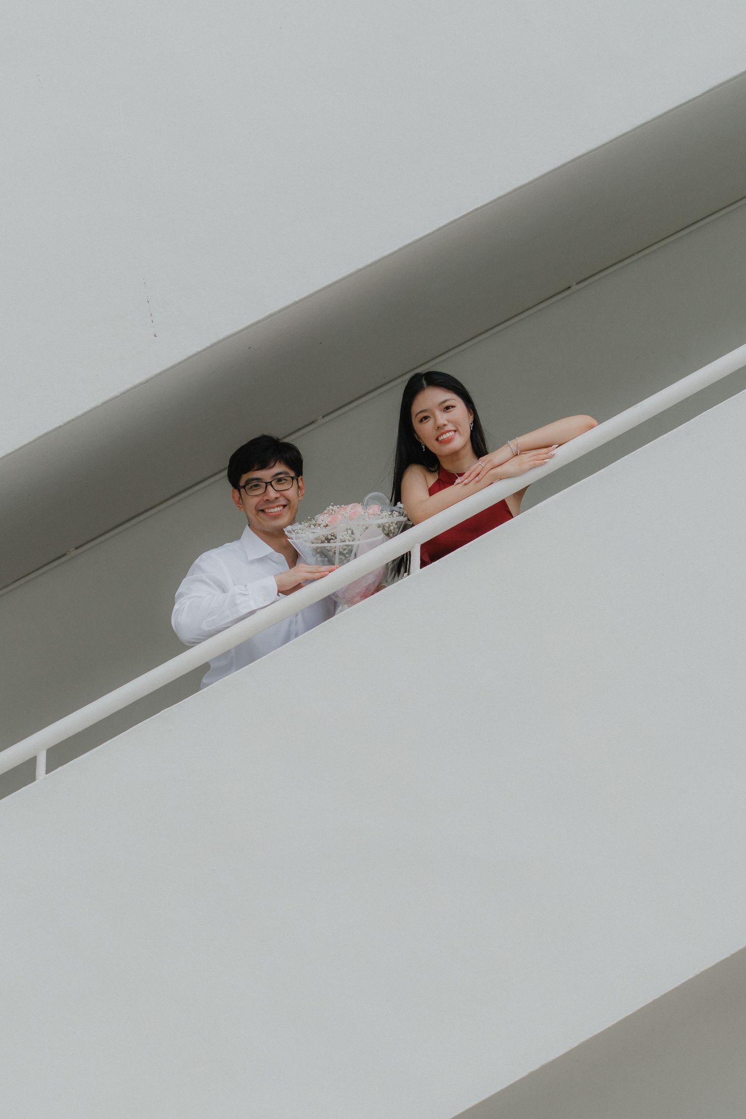 Two people smile and lean against a white railing on what appears to be a modern building's exterior balcony or walkway.