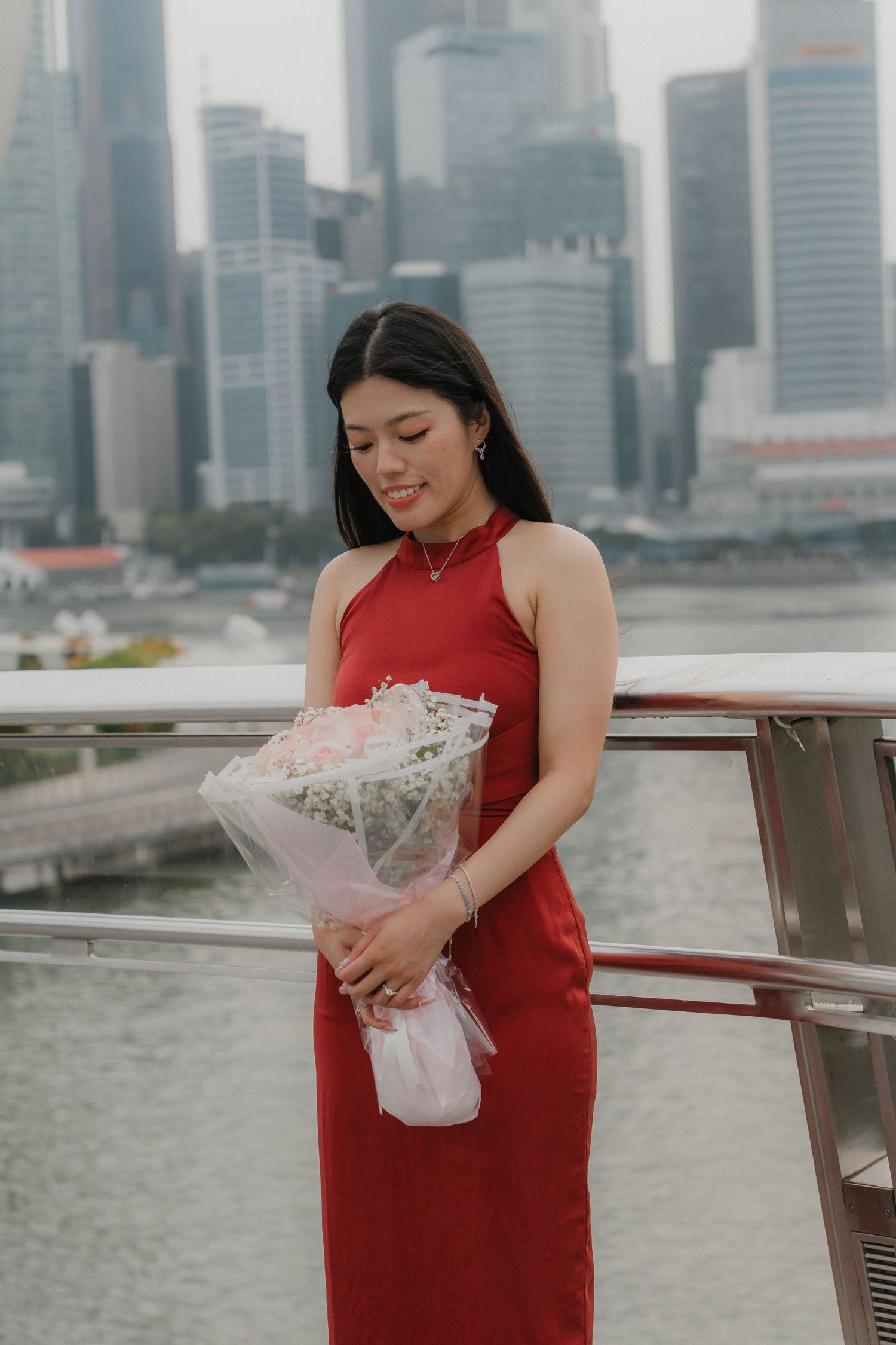 Woman in red dress holds white bouquet against Singapore city skyline.