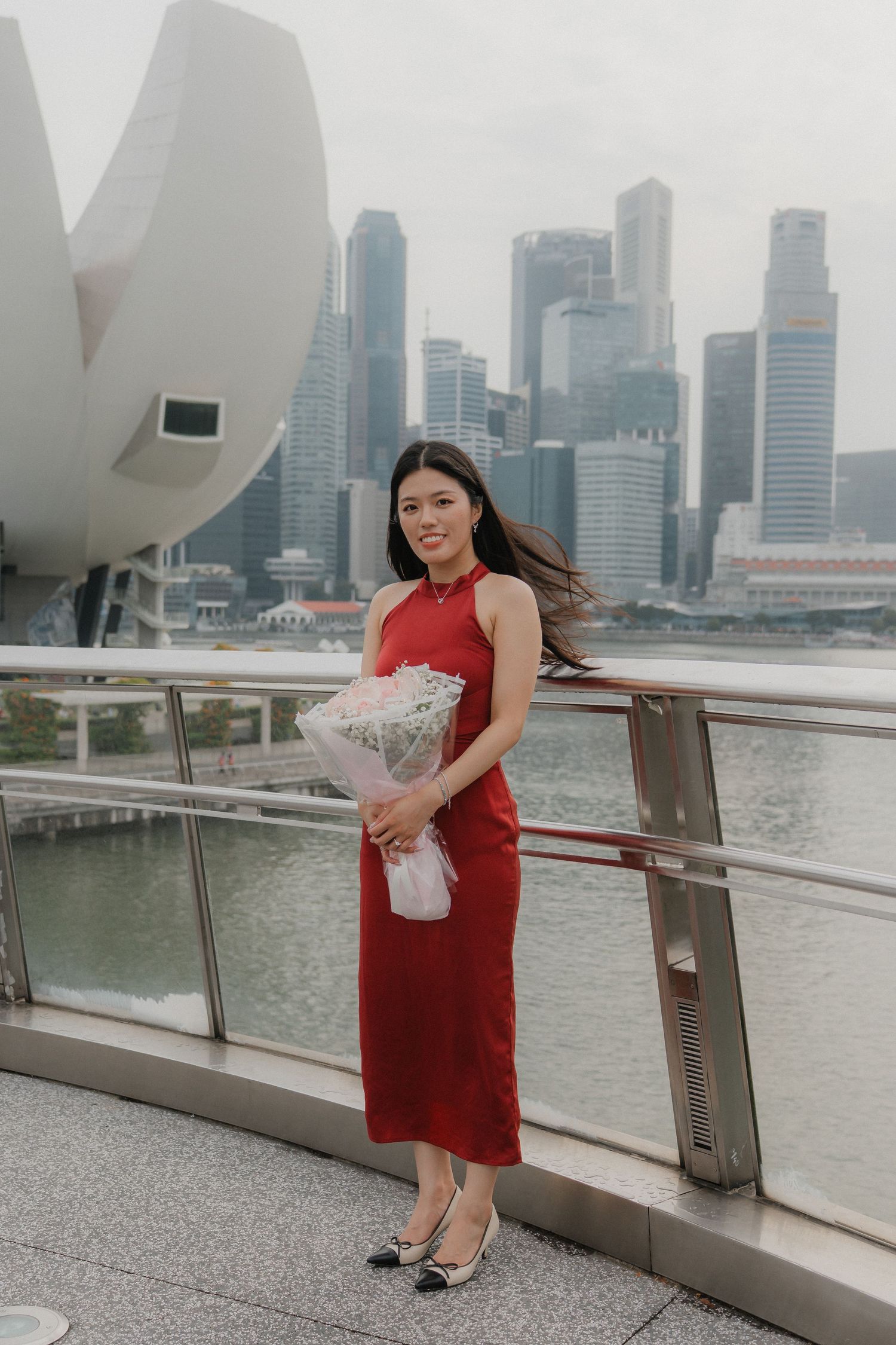 Red dress with bouquet against Singapore skyline backdrop at waterfront viewing area.
