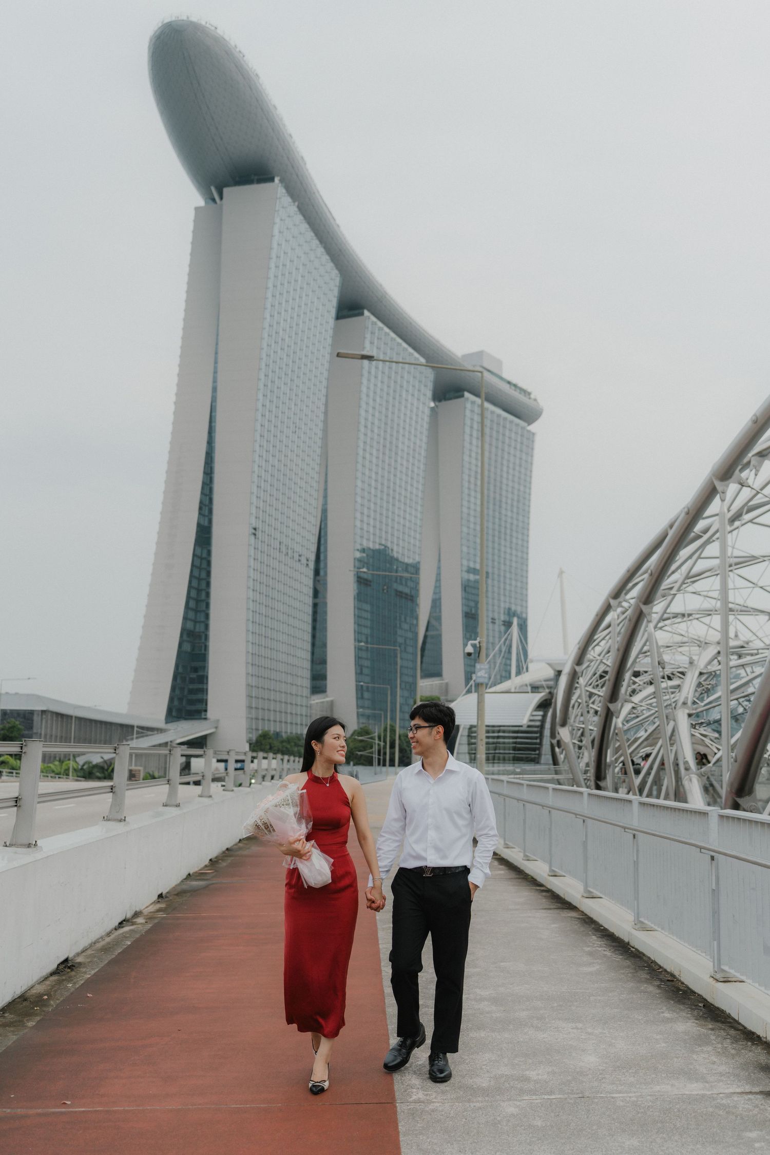 A couple walks along a waterfront bridge with Singapore's iconic Marina Bay Sands hotel in the background.