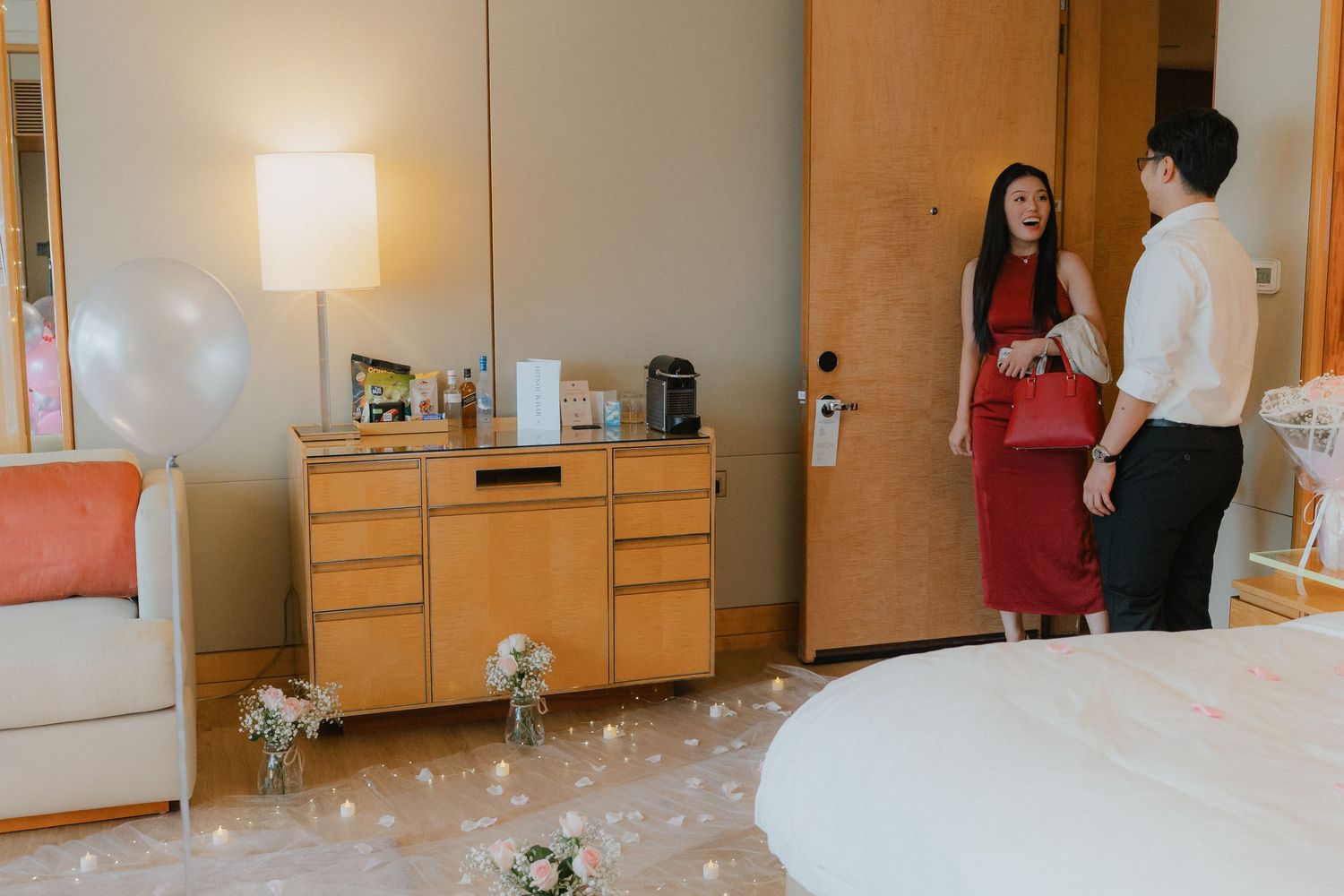 Two people having a conversation in a hotel room decorated with balloons and flowers for a romantic occasion.