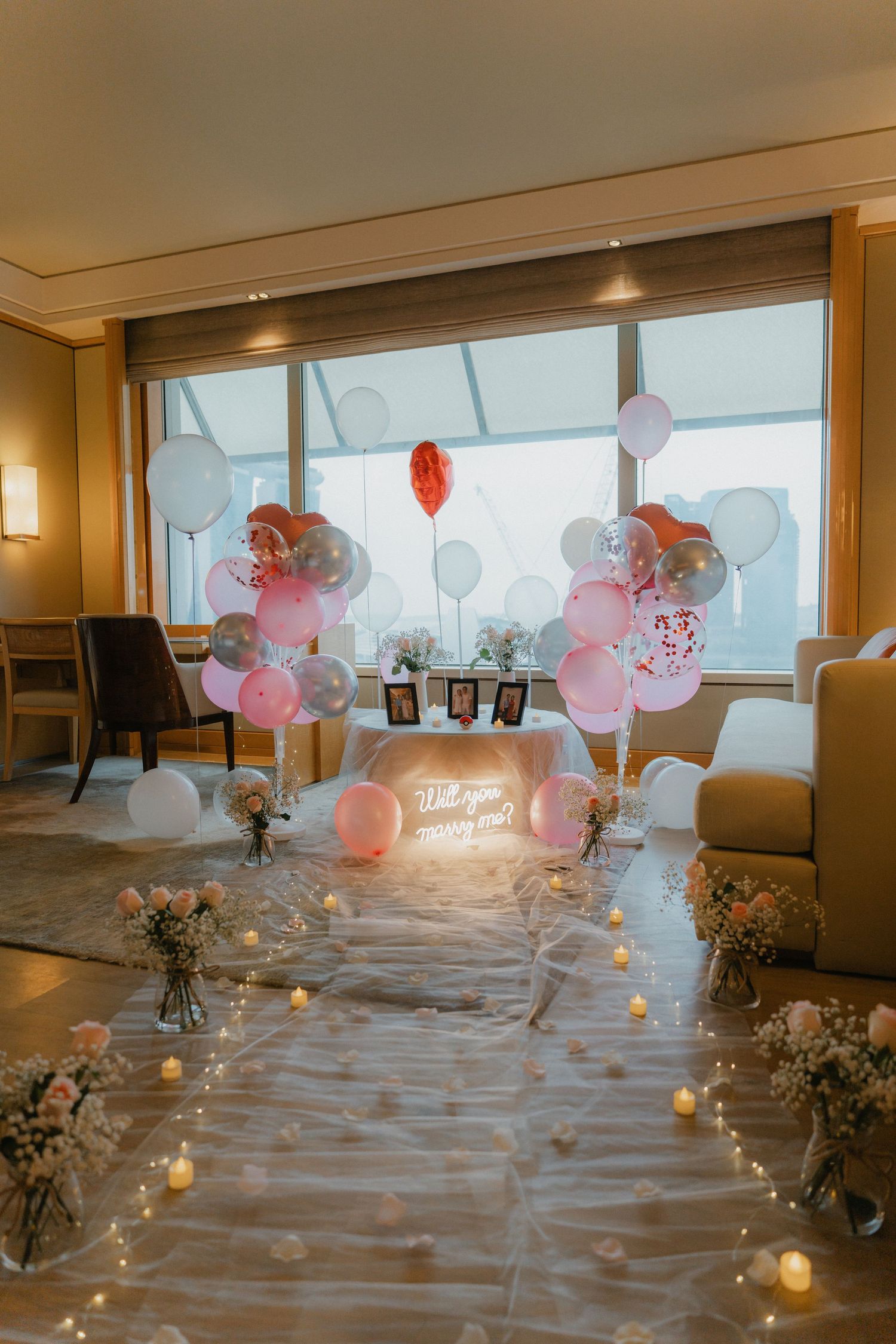 A romantic indoor proposal setup with pink and white balloons, fairy lights and floral decorations.