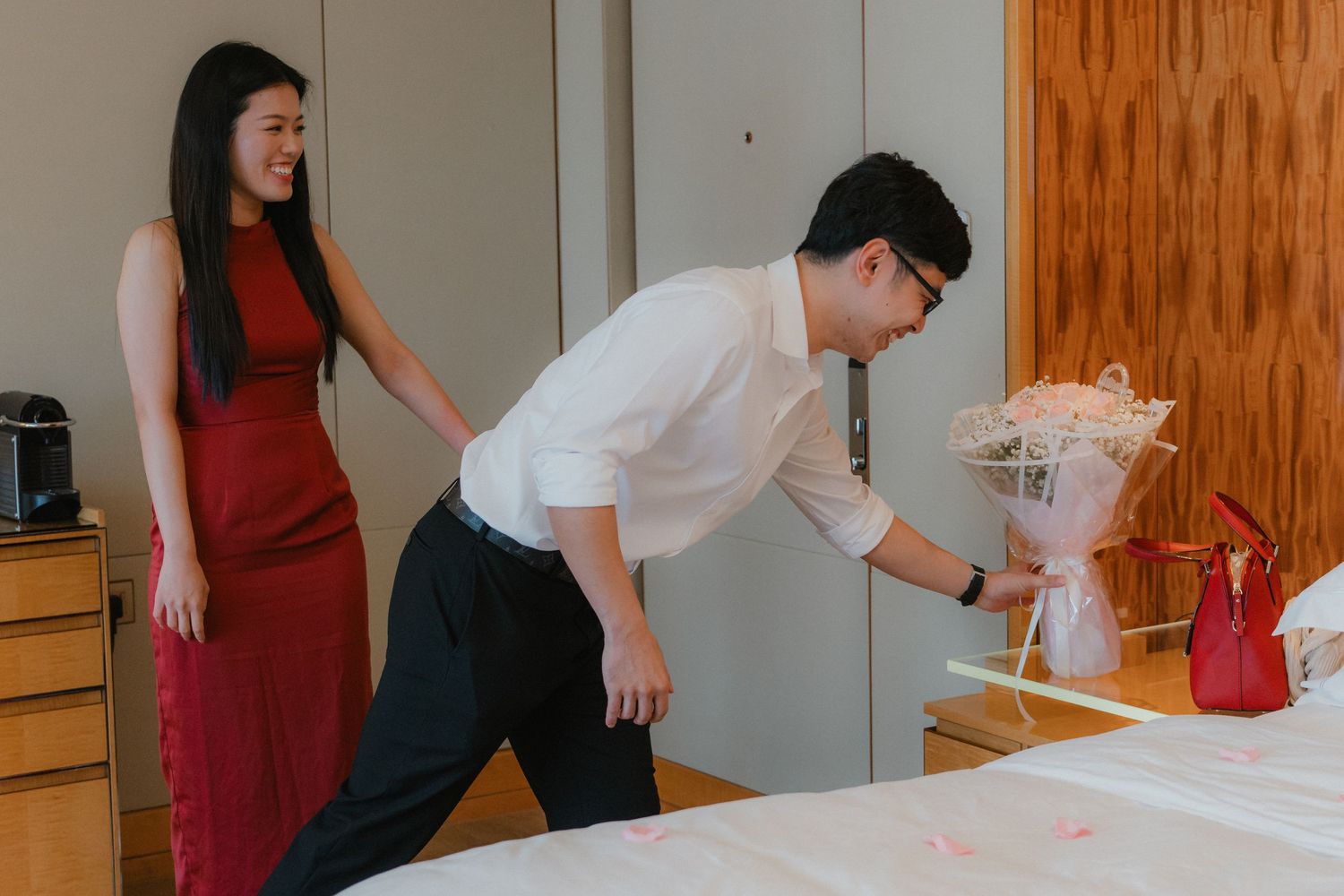 Two people in formal attire stand in a room, one wearing a red dress and another in white shirt leaning over a table.