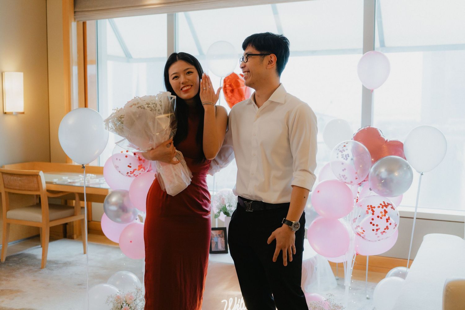 Couple celebrates in hotel room decorated with pink balloons and romantic decor.