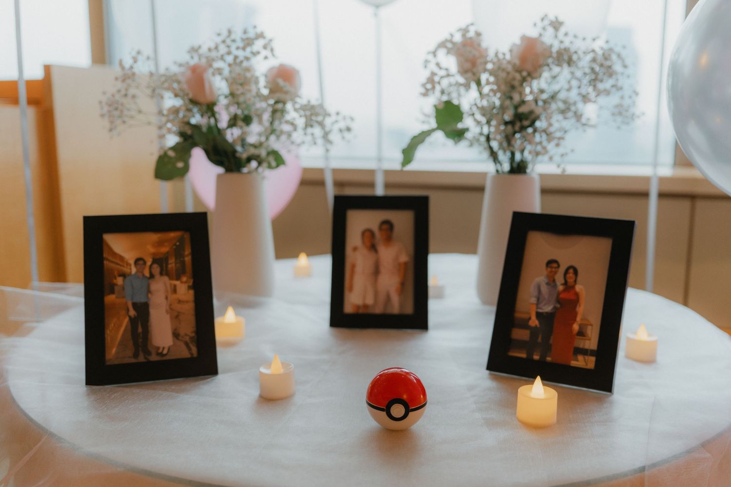 White floral arrangements with candles and framed photographs line a windowsill in a bright interior space.