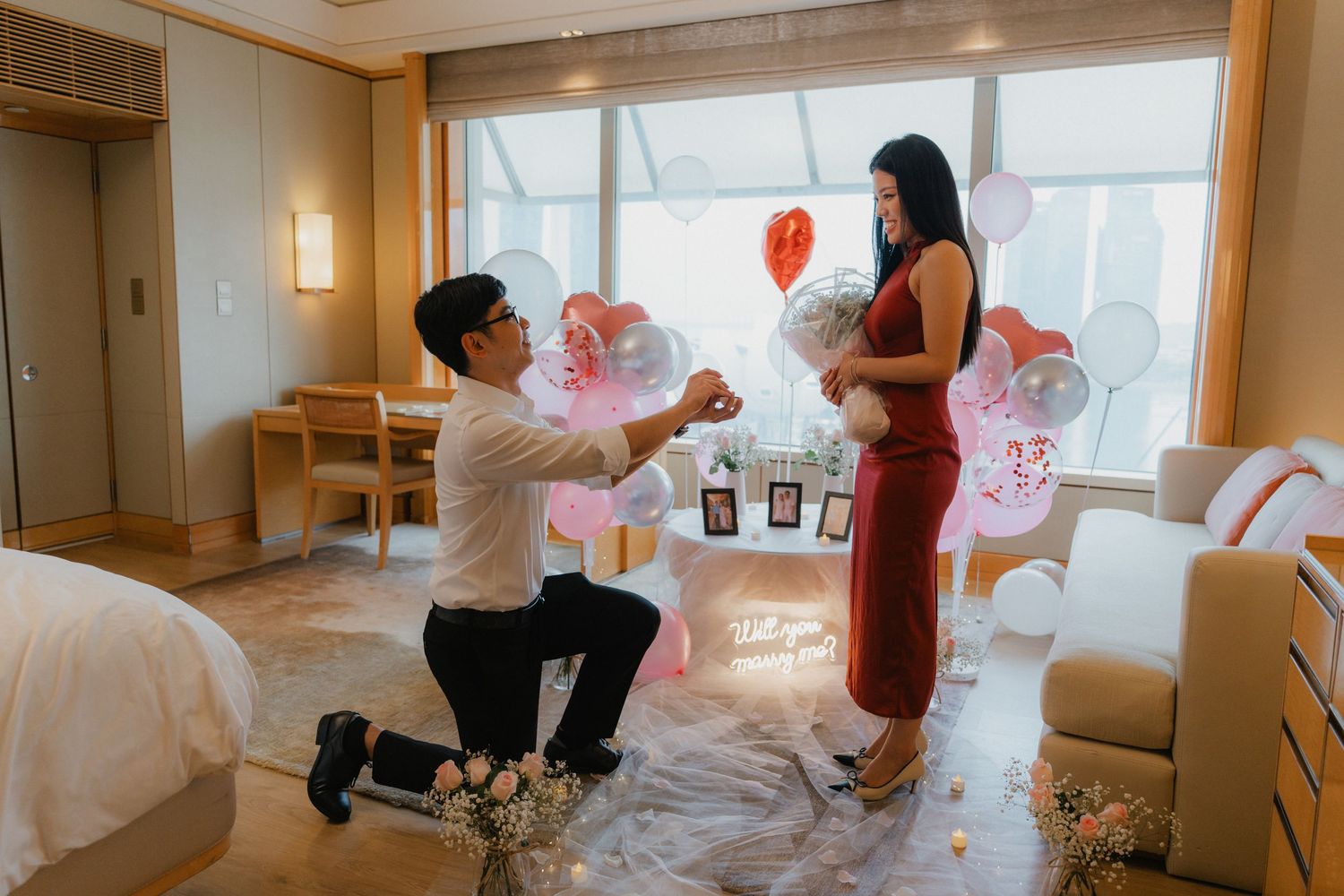 Pink and white balloons decorate a bright room with windows as people celebrate during a party.