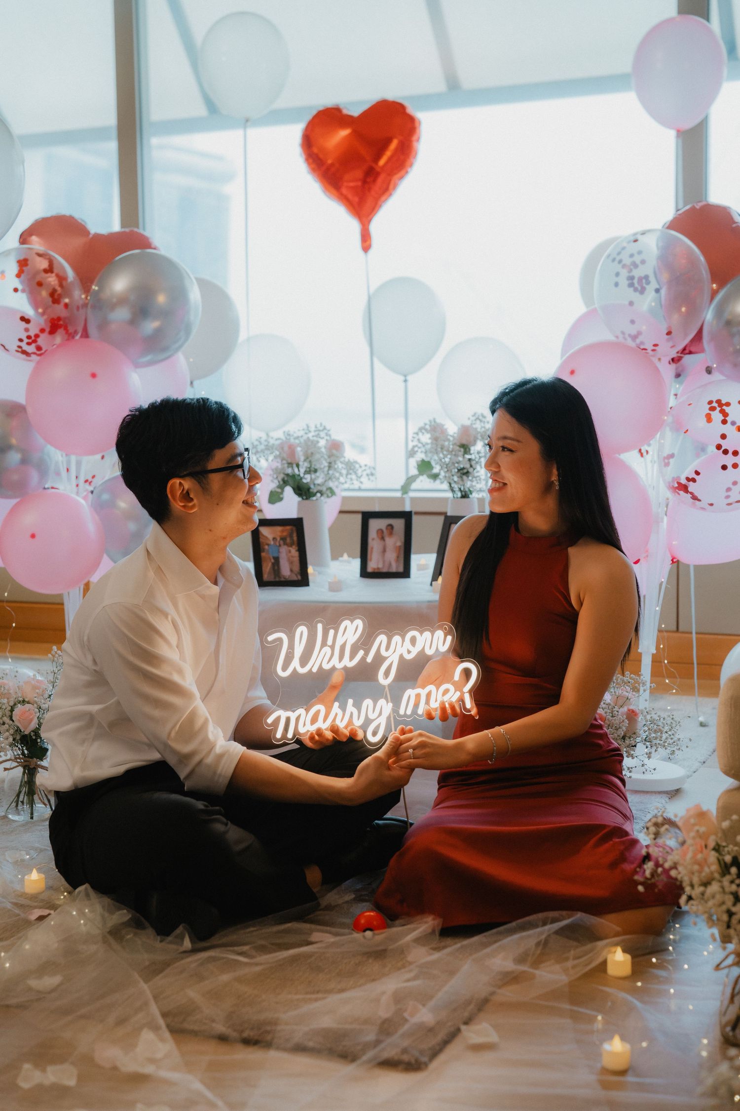 A romantic proposal setup with pink balloons and a neon 'Will you marry me' sign in a bright room.