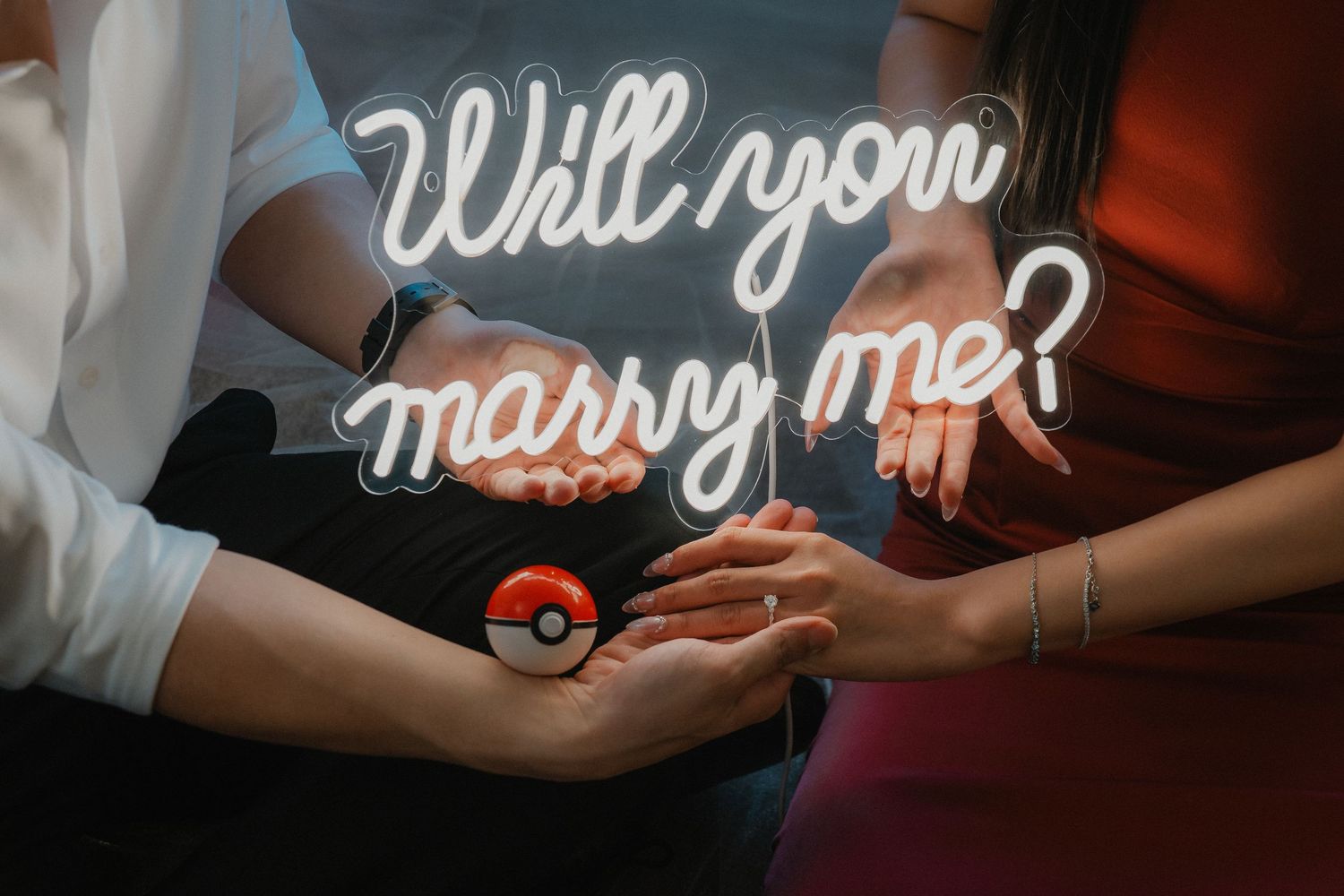 A close-up of hands holding a Pokemon-themed marriage proposal with illuminated neon 'Will you marry me?' signage.