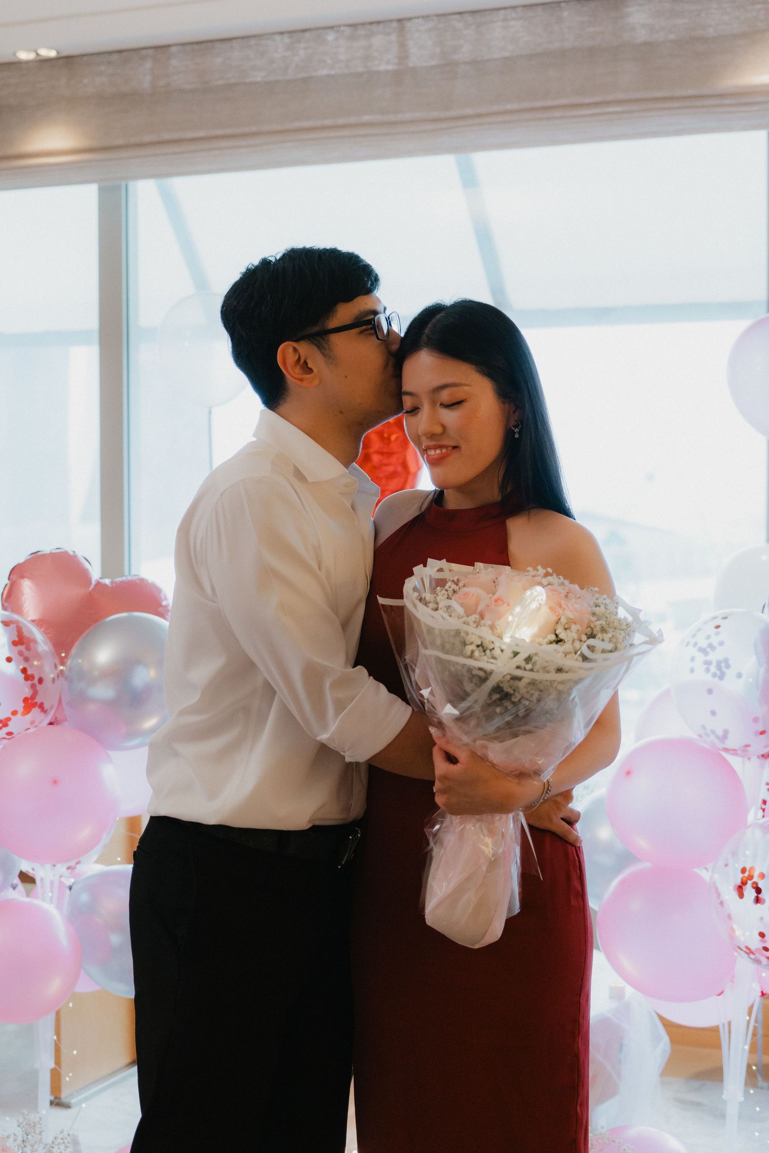 An intimate proposal moment surrounded by pink balloons and romantic decorations near large windows.