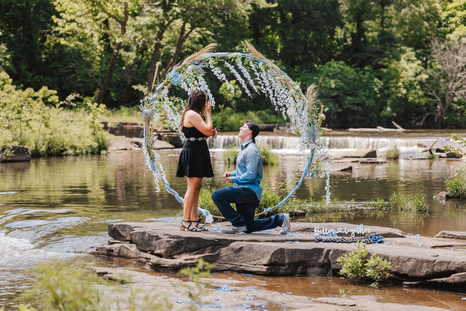 A romantic proposal moment on rocks by a scenic pond with a decorative arch in nature.