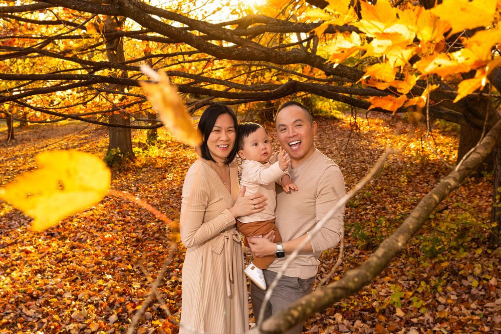 Family of three posing among golden autumn leaves in a sunlit forest, parents holding baby in warm fall colors.
