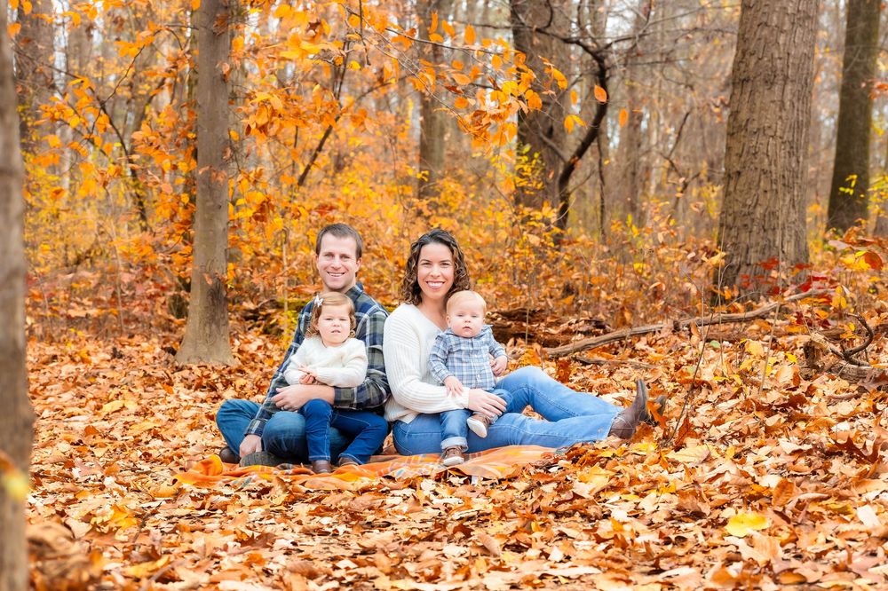 Family of four sitting in autumn leaves among trees with golden foliage in a forest setting.