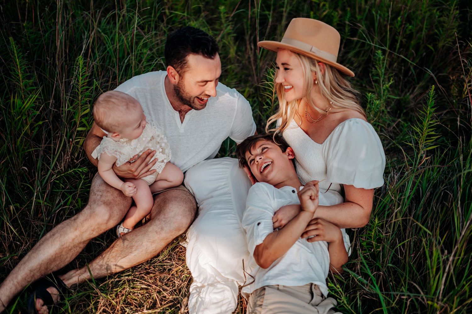 A family shares joyful laughter while sitting together outdoors surrounded by tall grass at sunset.