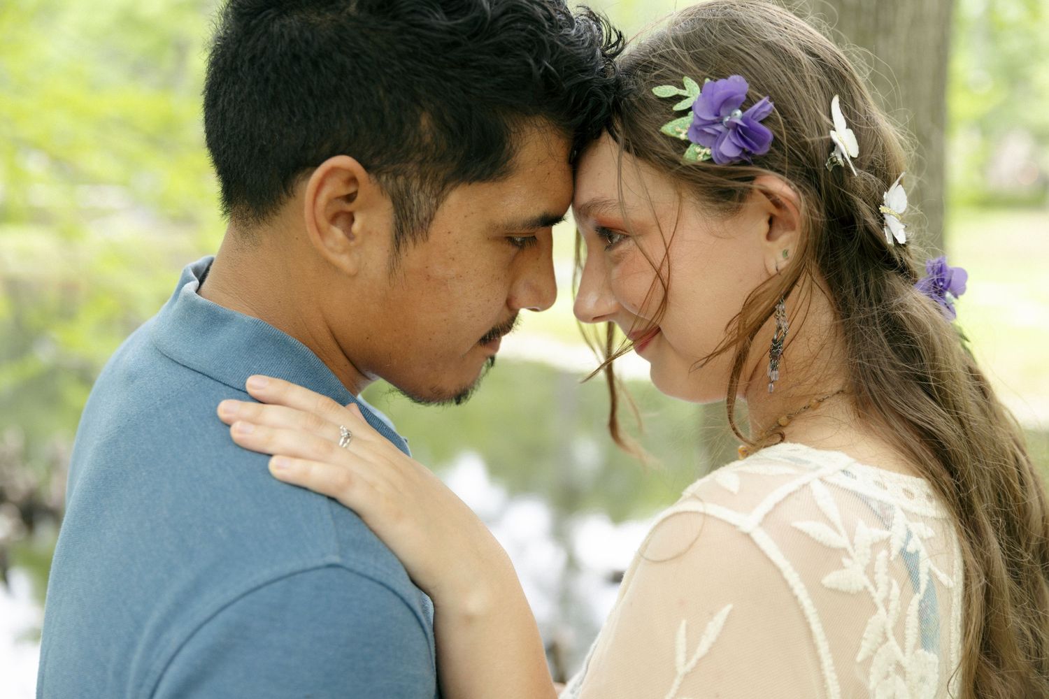 A tender moment between two people in a garden setting, with delicate purple flowers in her hair and a soft summer glow.