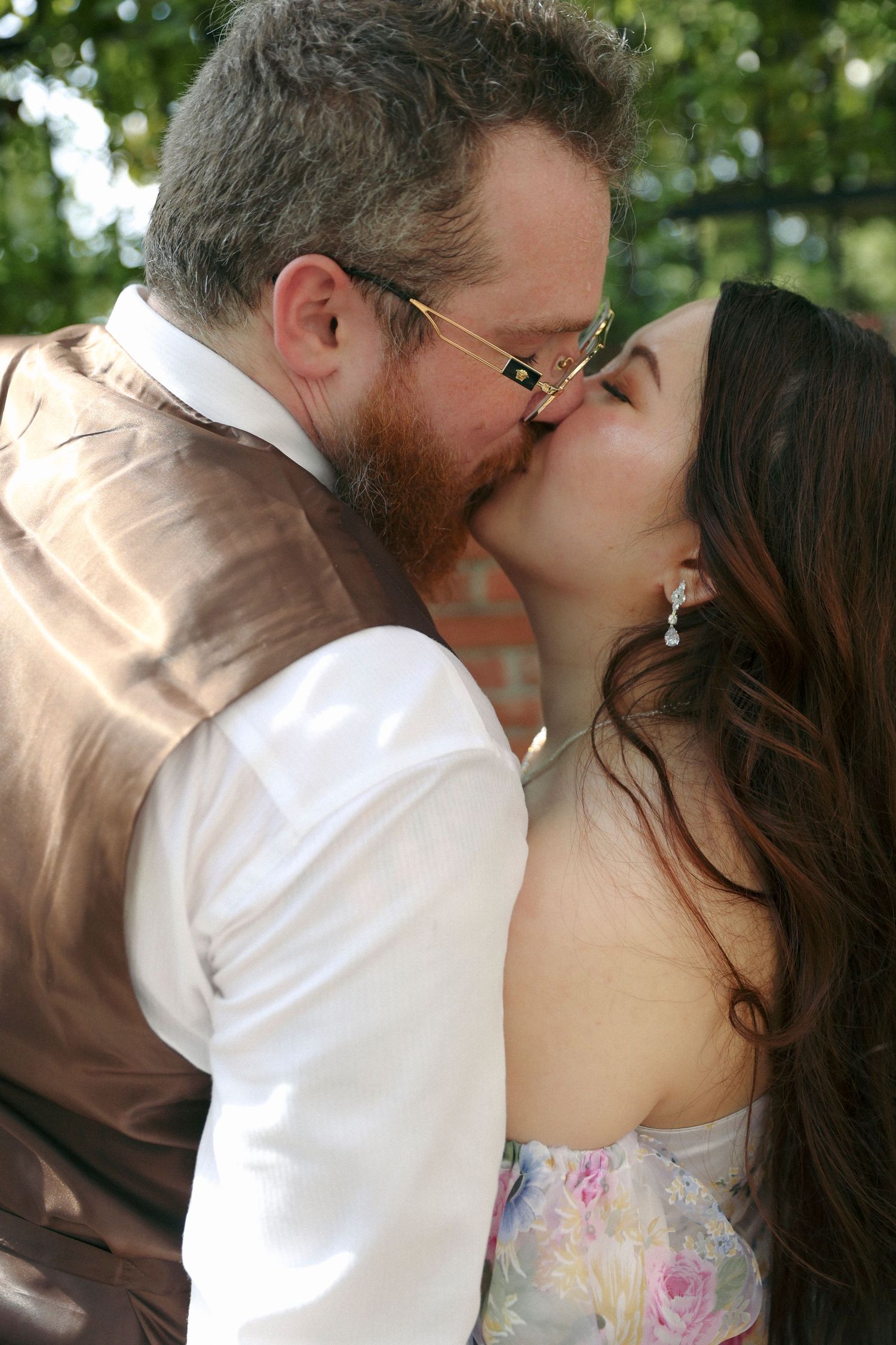 Two people share a romantic kiss outdoors, with the bride wearing a floral dress and groom in brown vest and white shirt.
