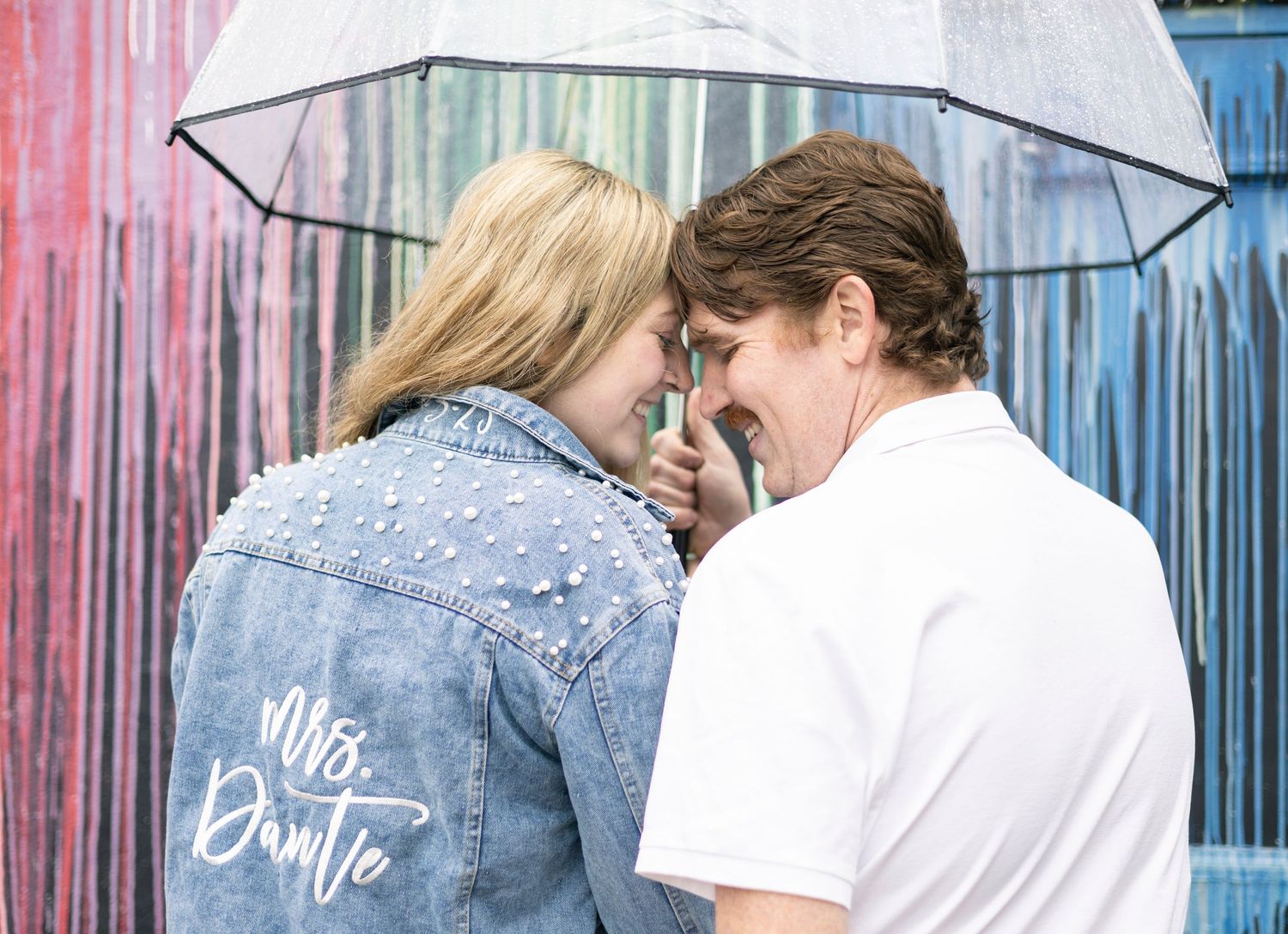 Three figures in denim and white clothing stand against a blue wall with a white umbrella.