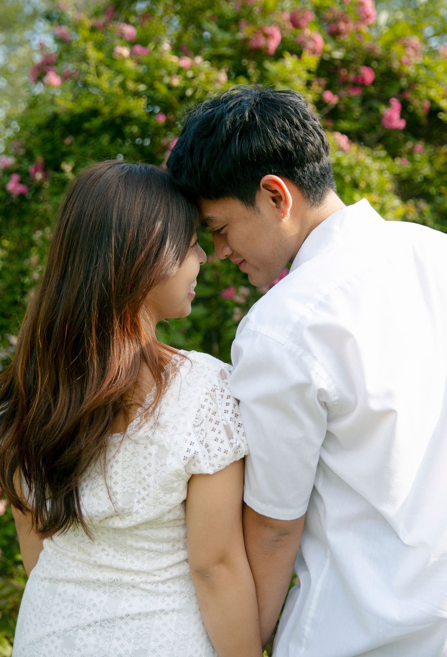 A romantic couple in white attire share tender moments against a backdrop of blooming pink roses in a garden setting.