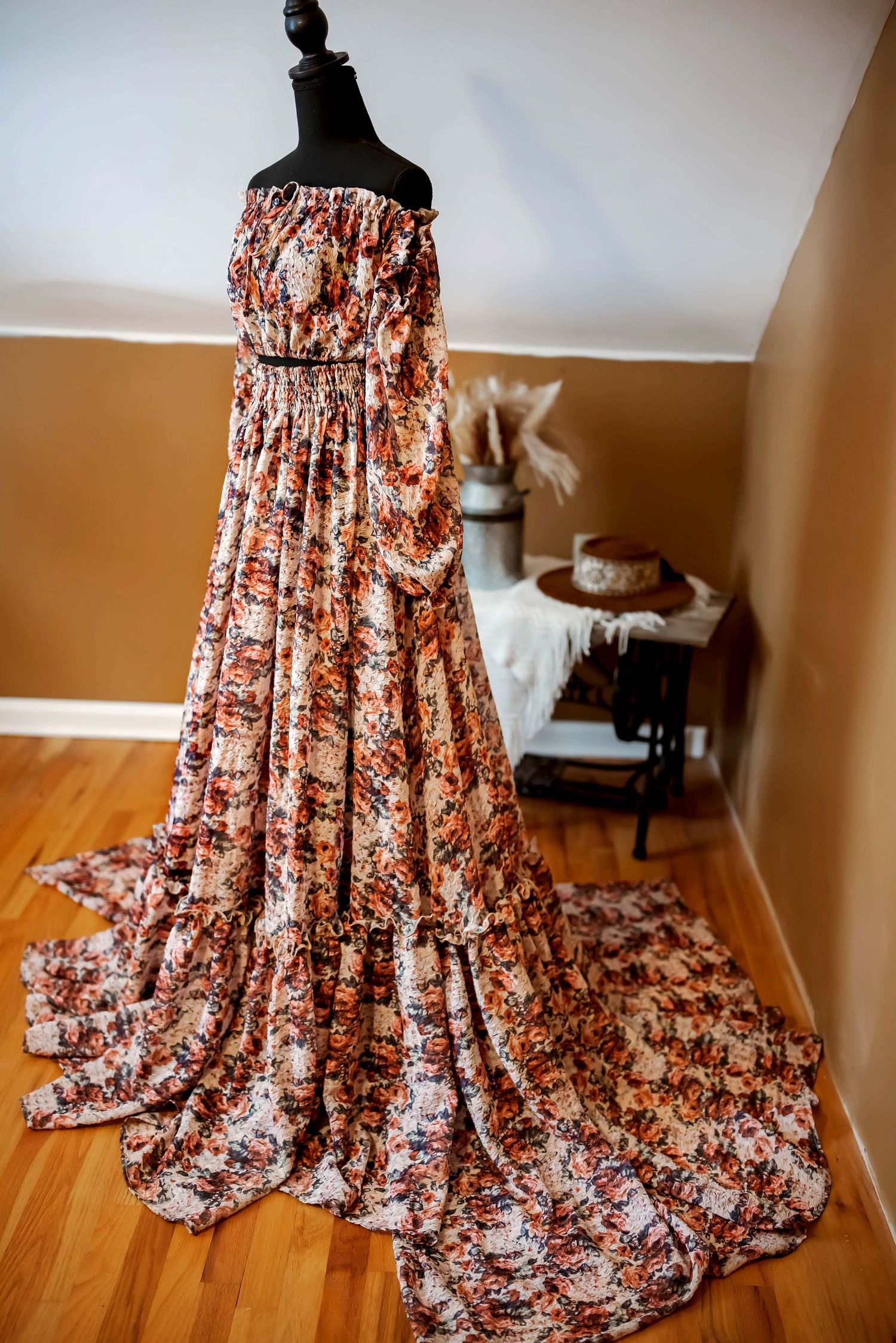 A teal and brown floral patterned maxi dress with off-shoulder sleeves displayed on a black mannequin against hardwood flooring.