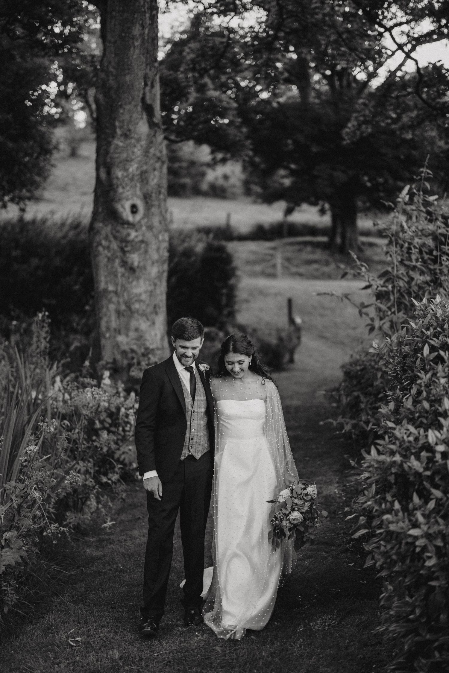 A couple stands together on a garden path surrounded by lush greenery in this black and white wedding photo.