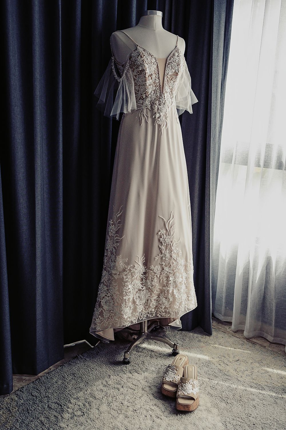 An elegant white wedding dress with delicate lace details and flowing sleeves displayed on a mannequin with shoes below.