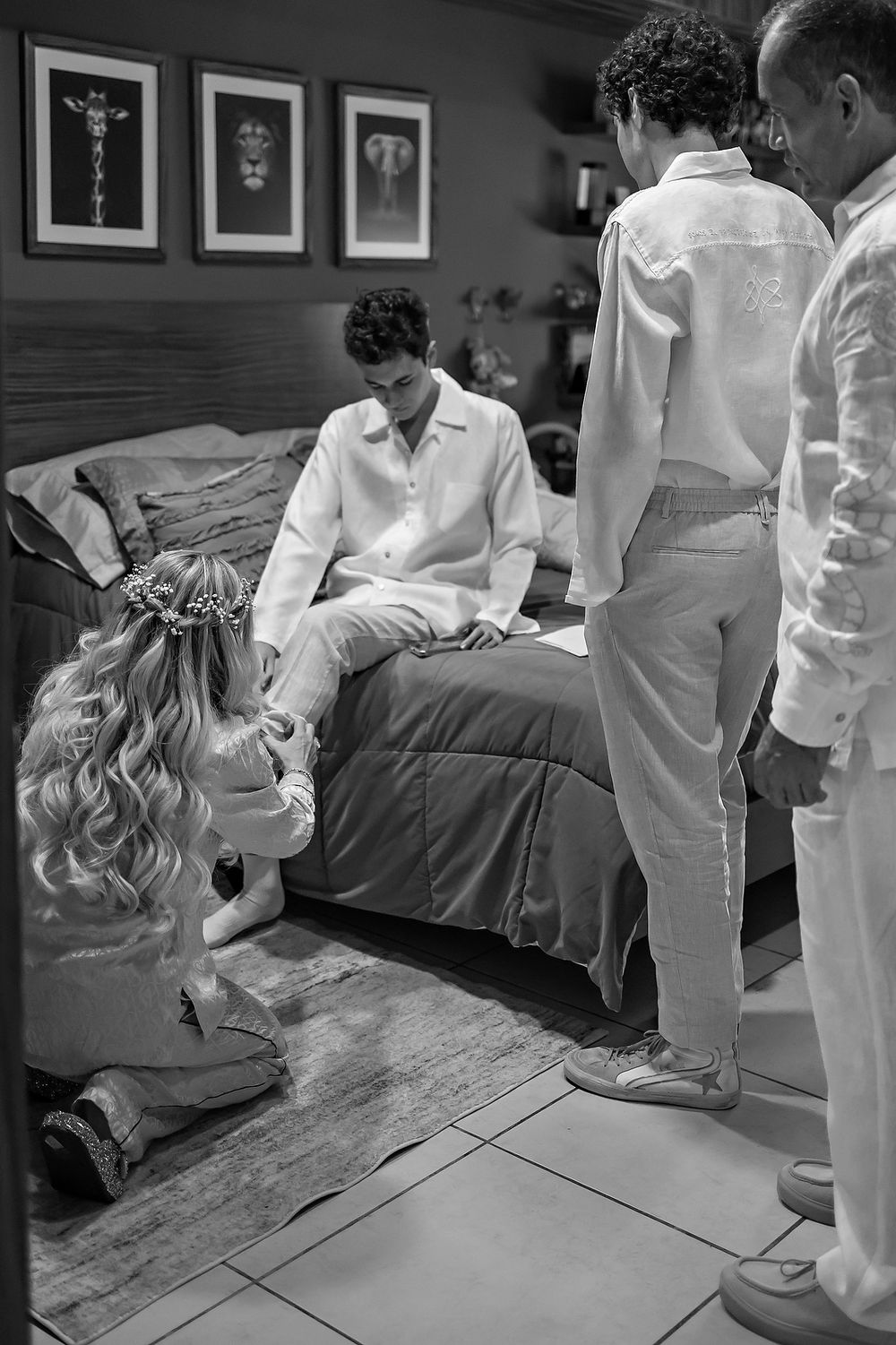 Black and white photo of people getting ready in a bedroom with three framed artworks hanging on the wall above.