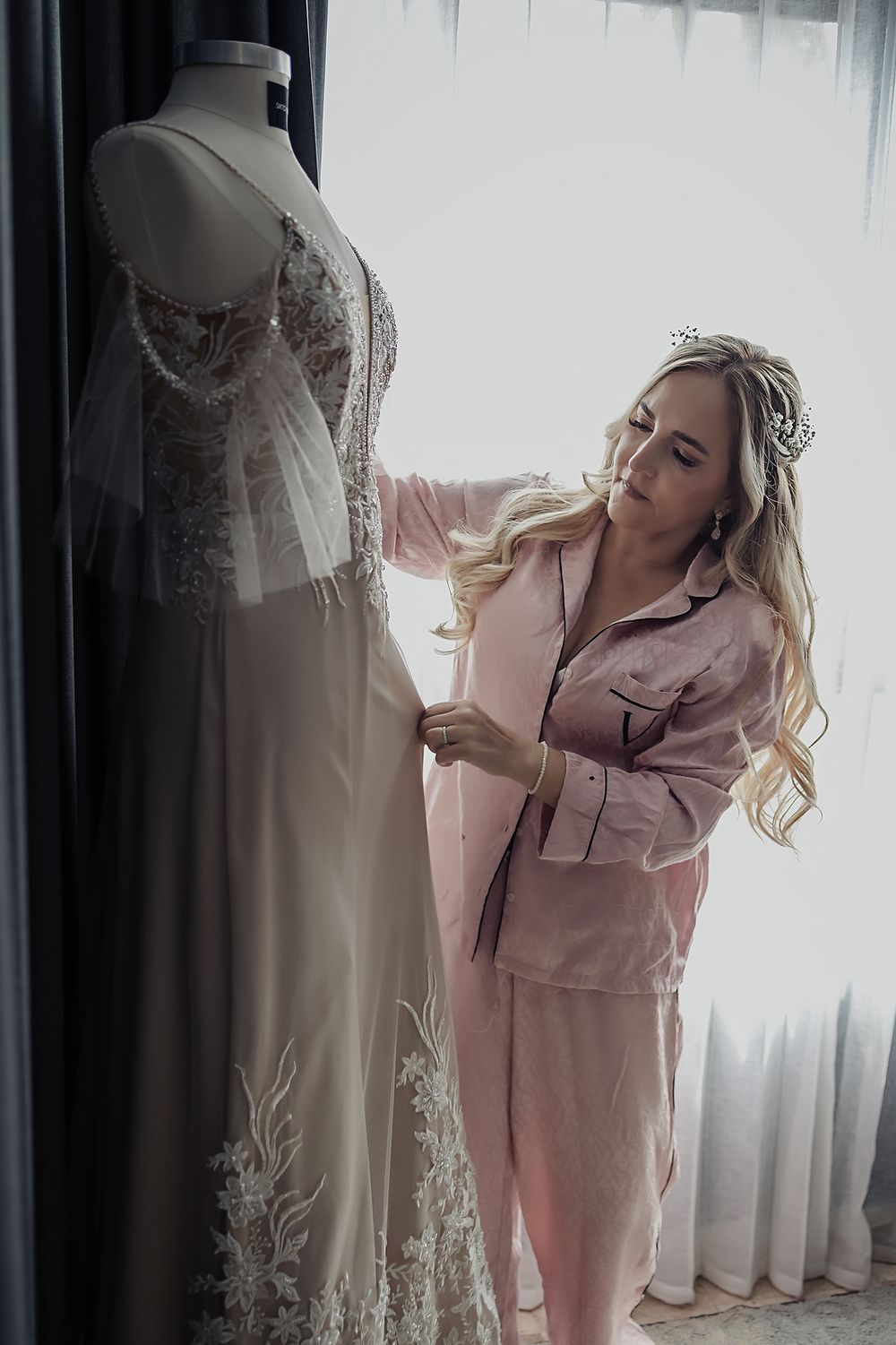 A woman in pink satin pajamas inspects a detailed lace wedding dress on a mannequin form near a window.