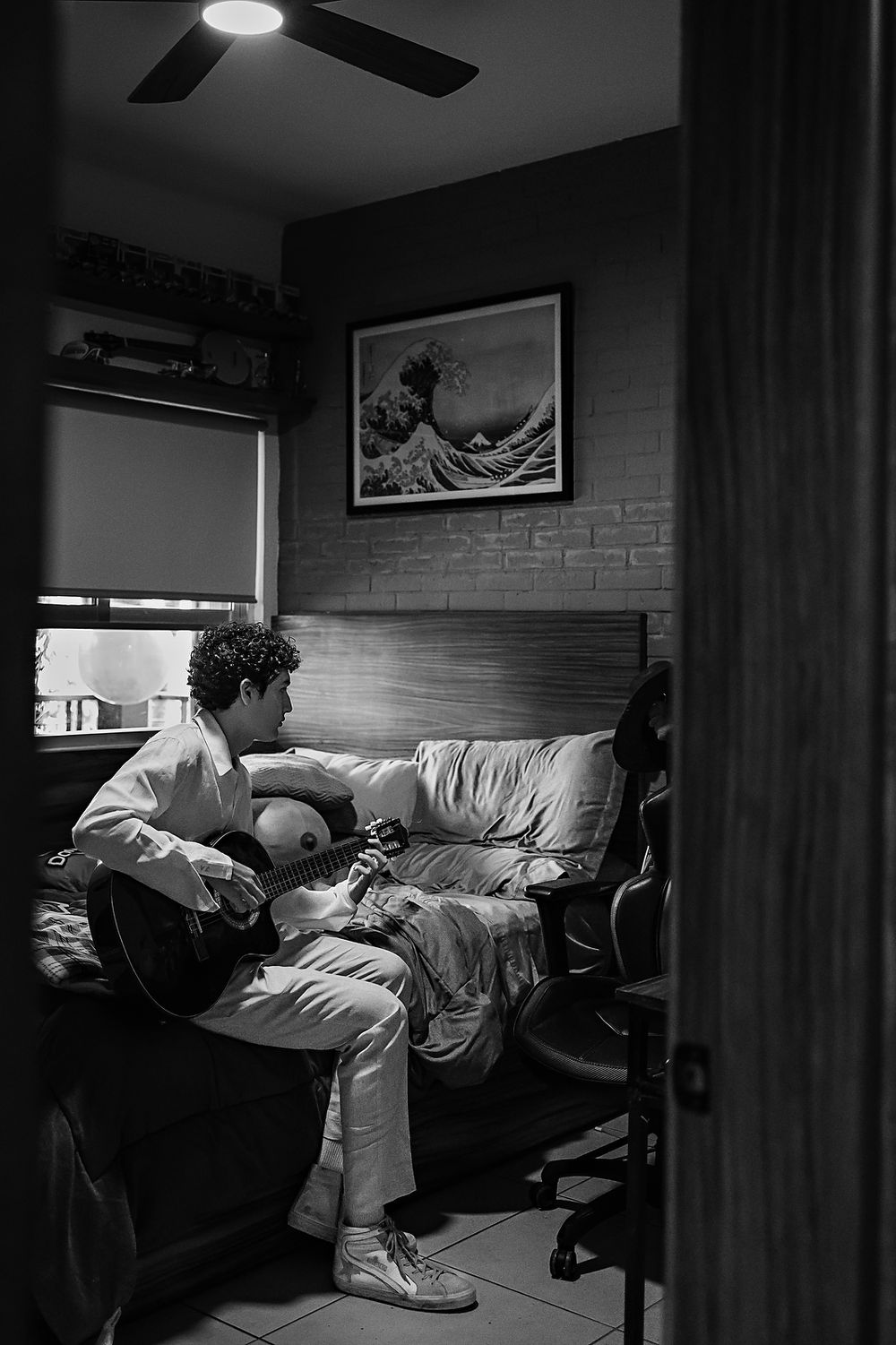 A black and white photograph of someone playing guitar in a cozy room with a framed wave artwork on the wall.