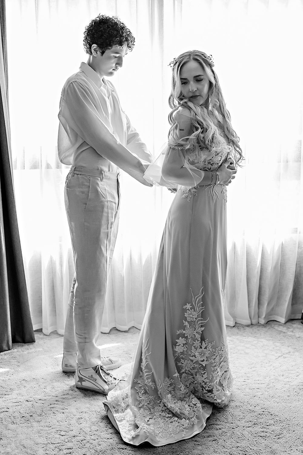 A young child and someone in a flowing dress dance together in a sunlit room with white curtains.