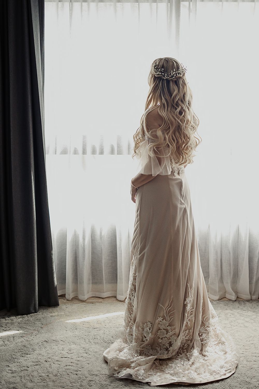 A bride stands in flowing white dress with lace details and long wavy blonde hair adorned with a delicate floral crown.