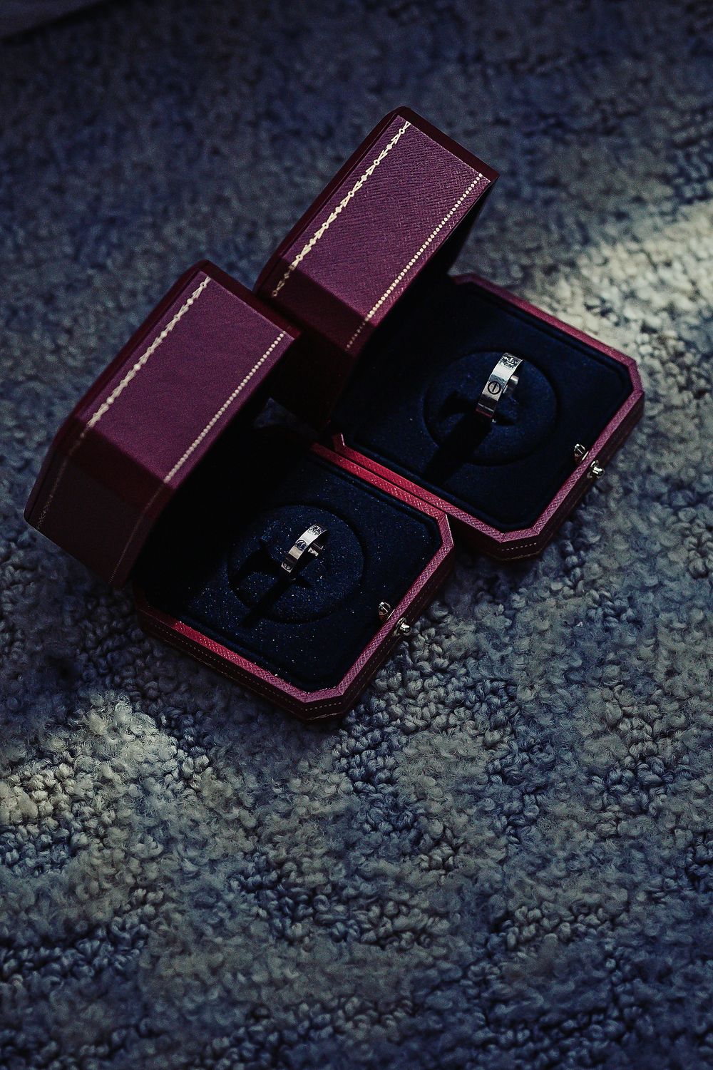 Two burgundy wedding ring boxes with sparkling engagement rings nestled in dark blue velvet interiors.