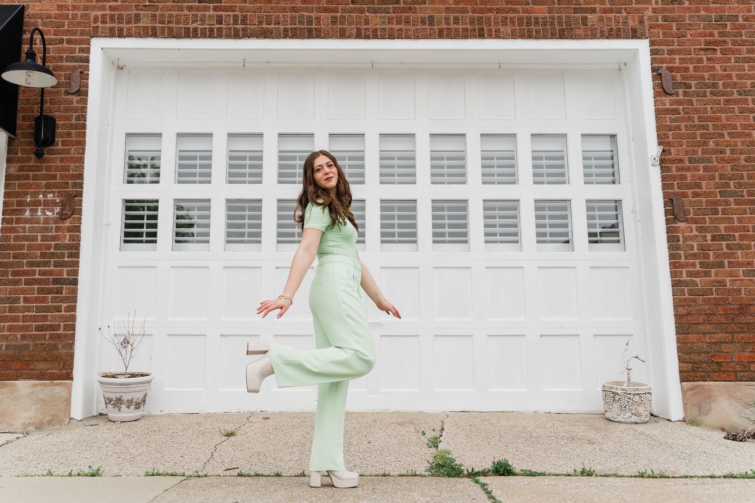 A joyful figure in mint green attire dances and leaps in front of a large white garage door and brick wall.